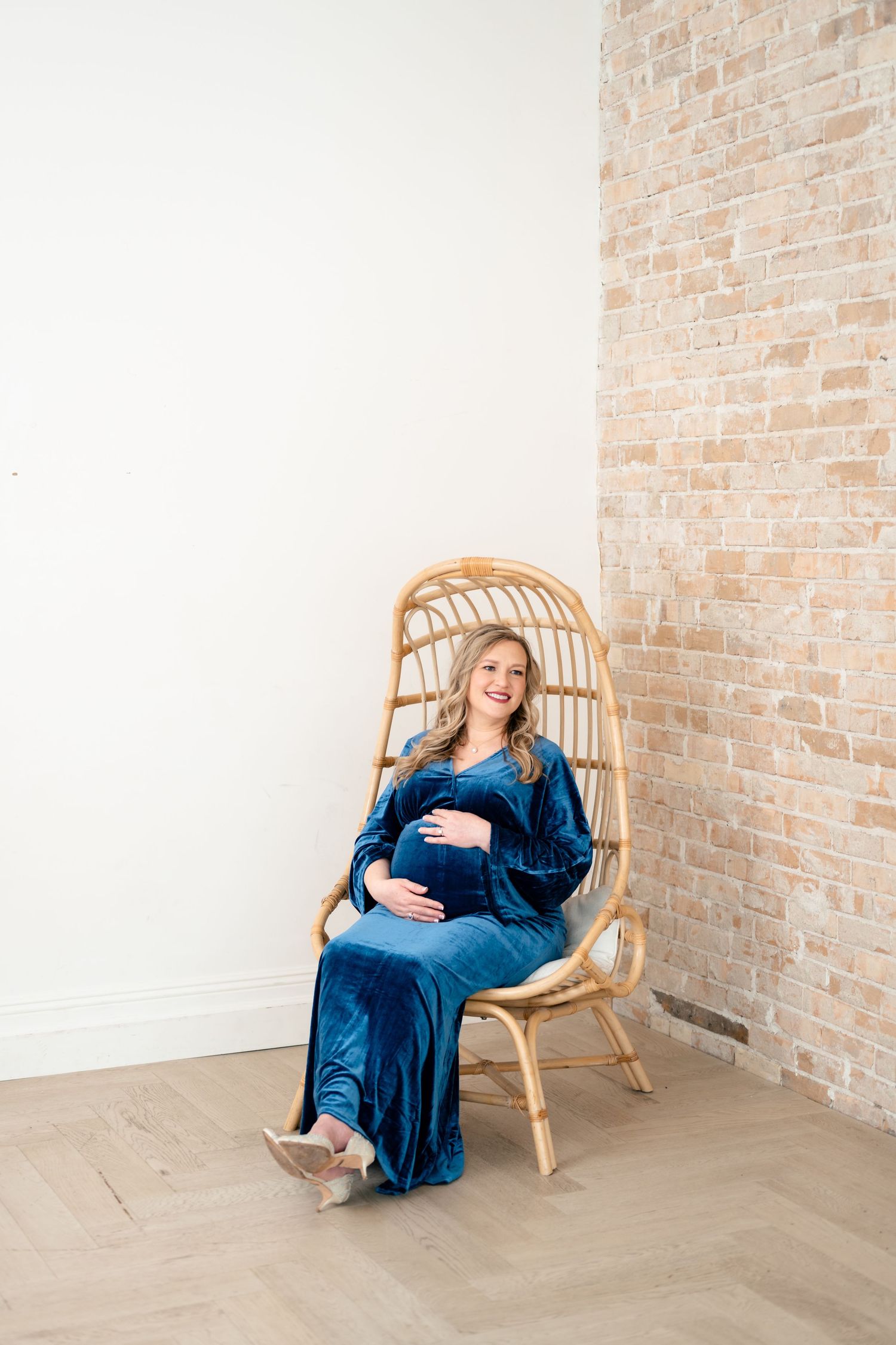Pregnant woman in blue velvet dress sitting in rattan chair, smiling and cradling baby bump near brick wall.