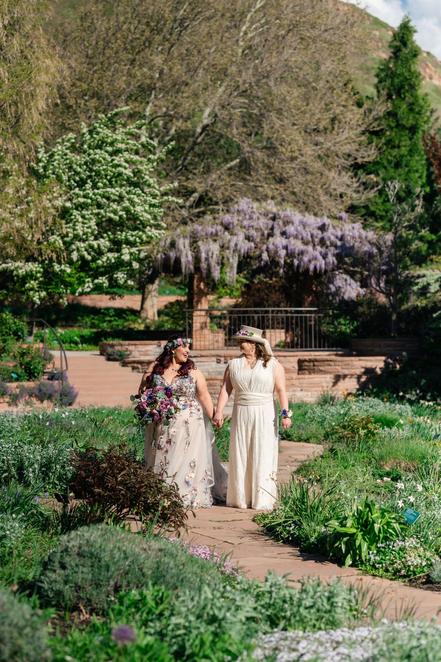 Two brides walk hand-in-hand through a lush garden path with blooming wisteria and white trees in spring.