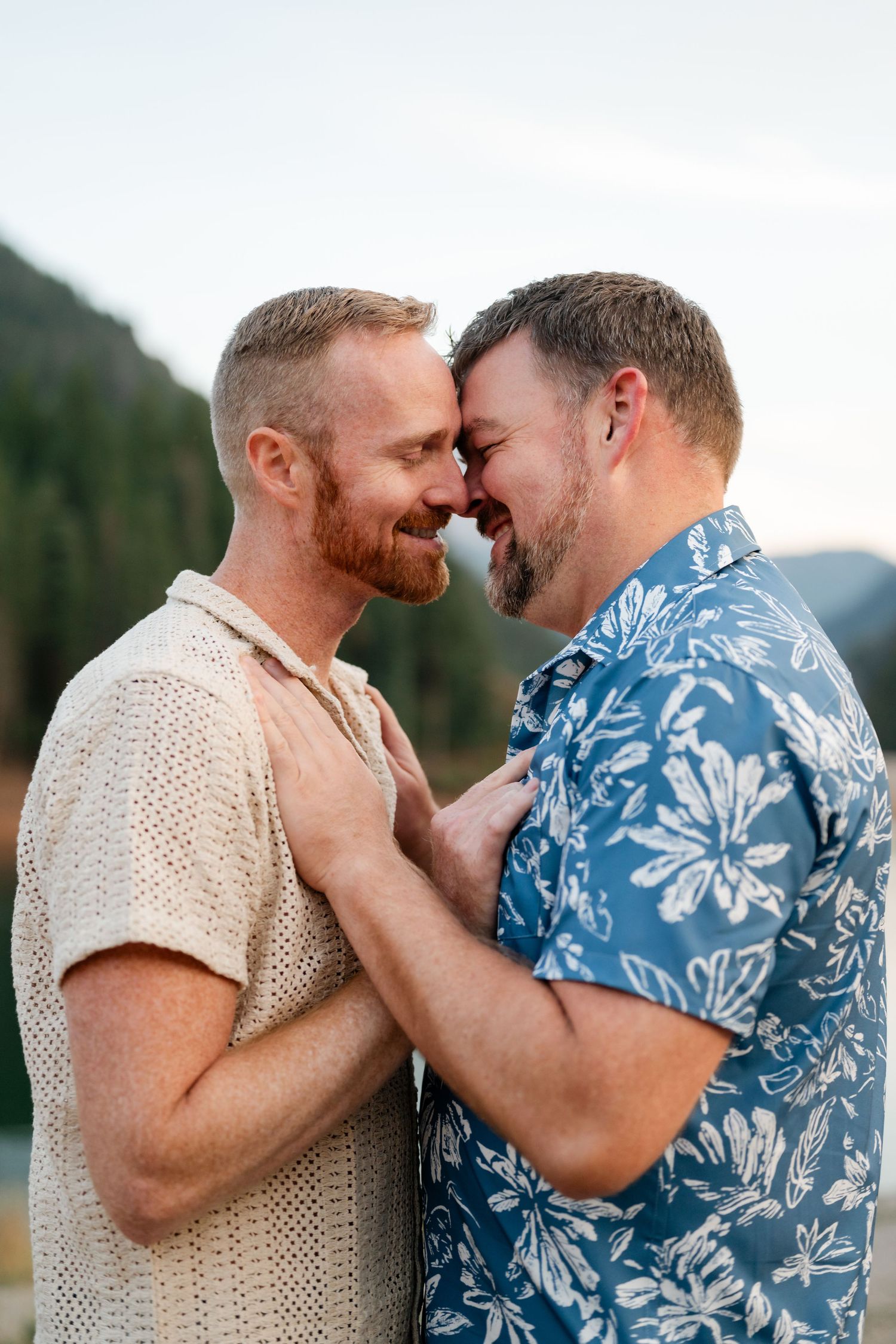 Two men share a romantic forehead touch while smiling outdoors near a mountain lake.