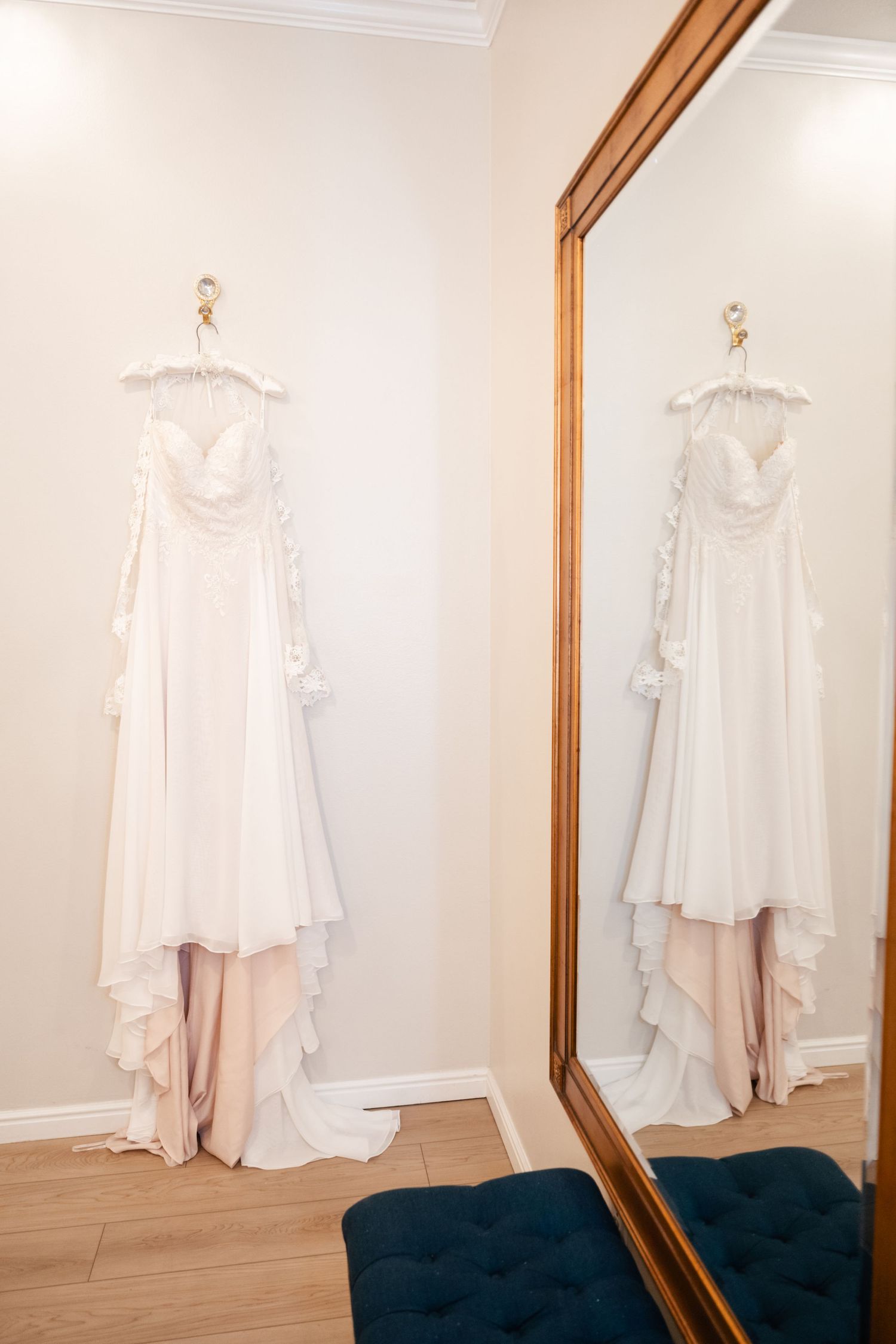 A white wedding dress with flowy details hanging on a hanger, reflected in a large mirror with a wooden frame.