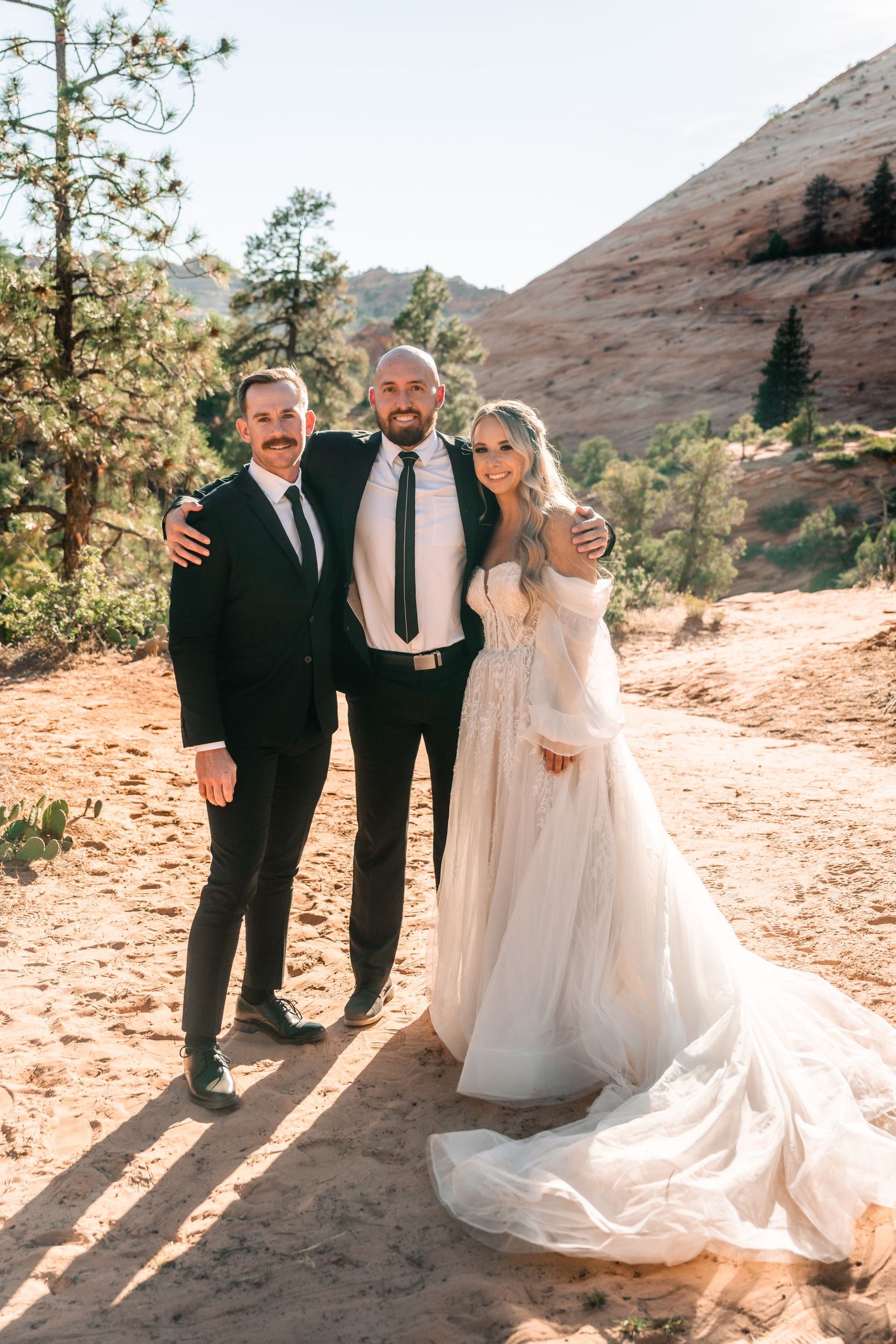 Three people pose together during a wedding celebration on a scenic mountain trail.