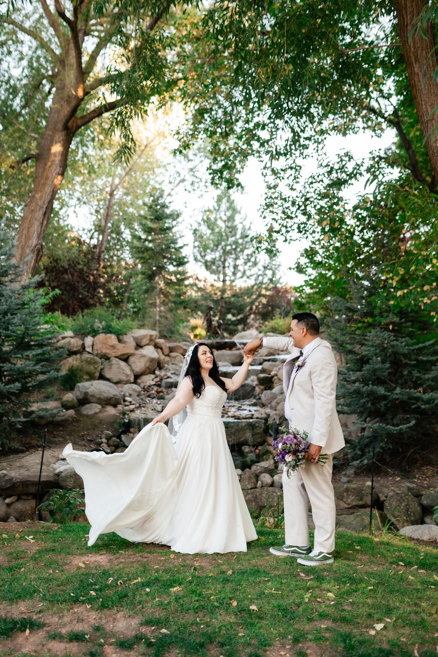 A couple shares a romantic moment in a garden setting with evergreen trees and a rock wall as their wedding backdrop.