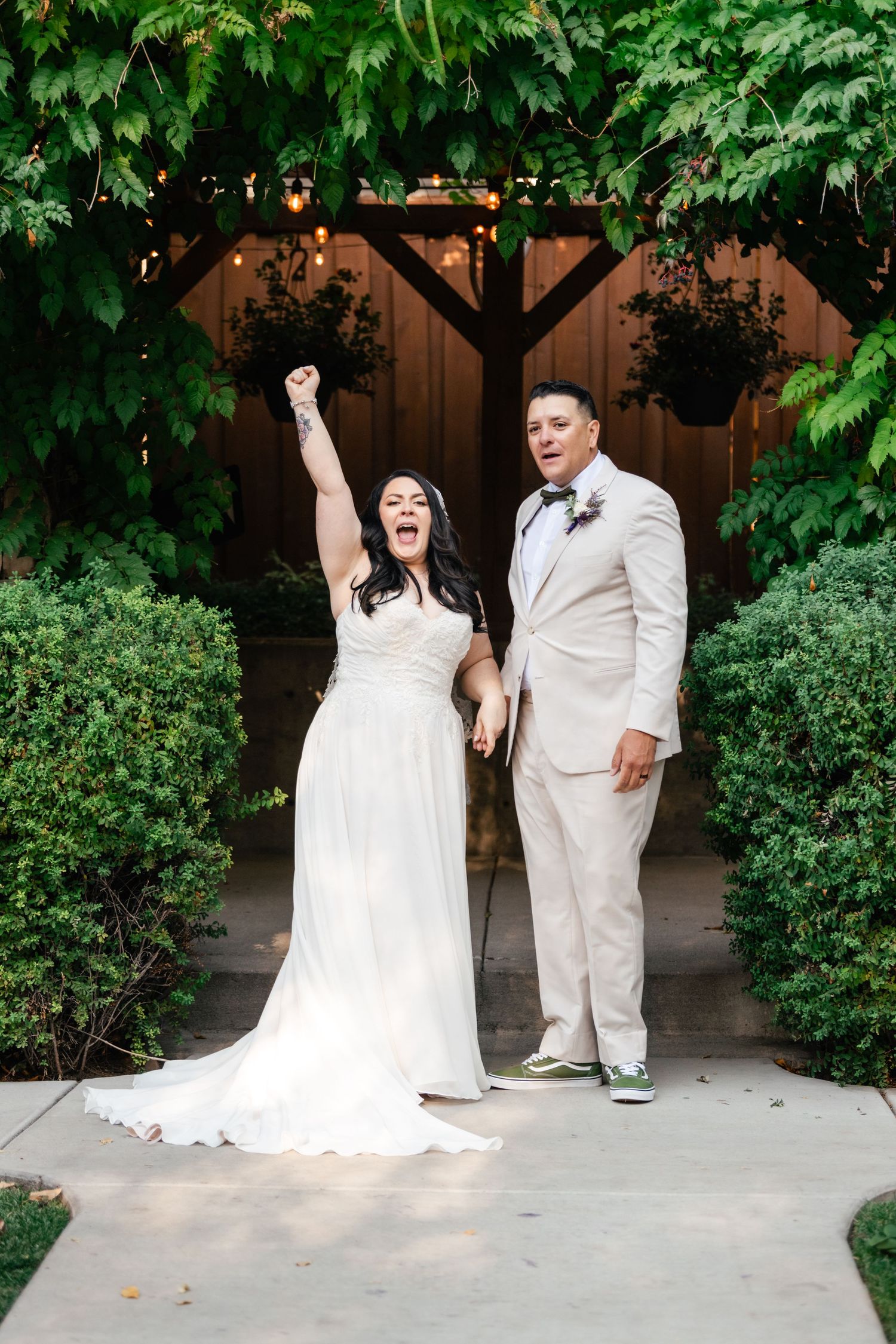 A celebratory wedding couple in white attire pose together on a garden path framed by lush greenery and a wooden arch.