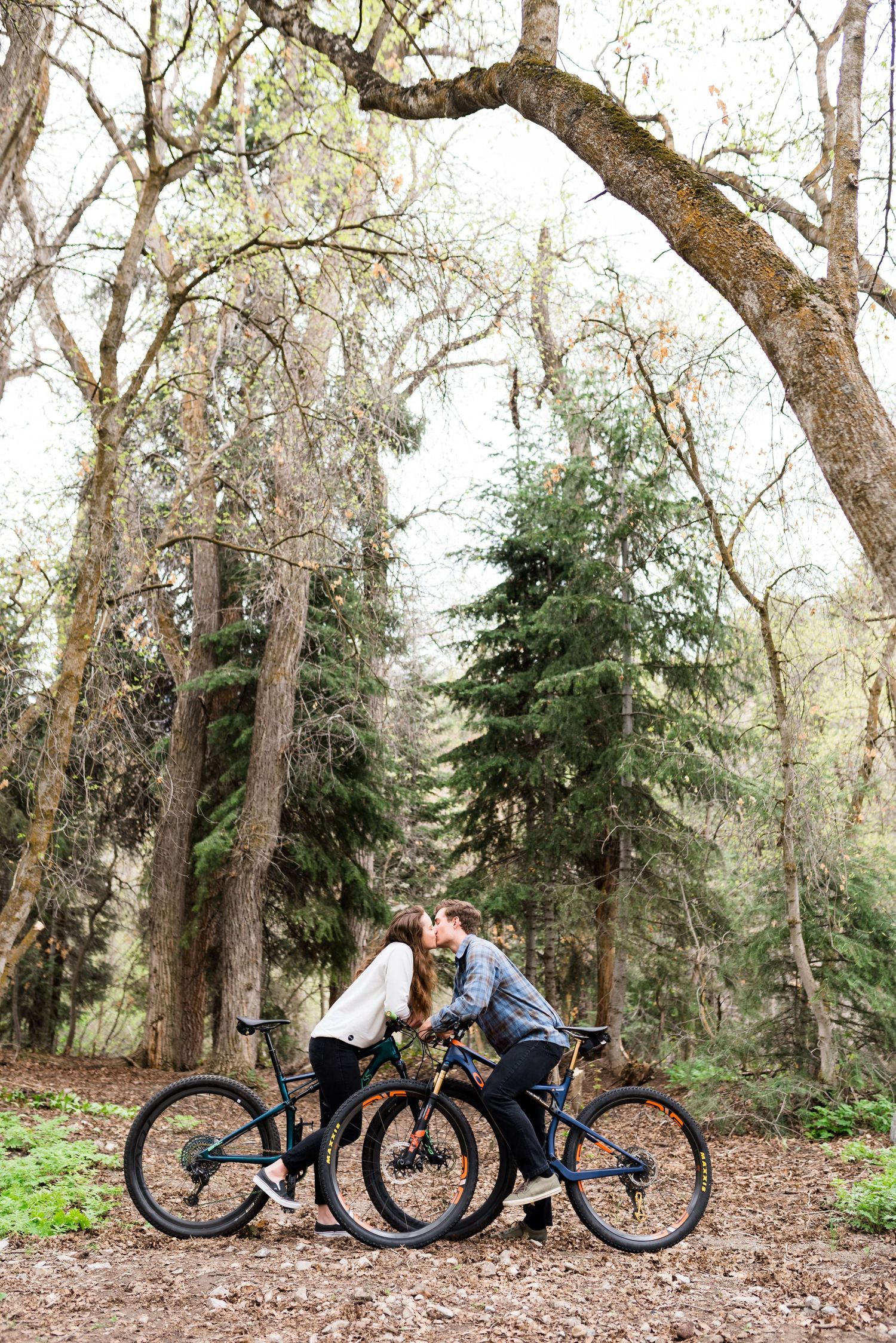 Couple sharing a kiss while leaning on mountain bikes in a lush forest setting.