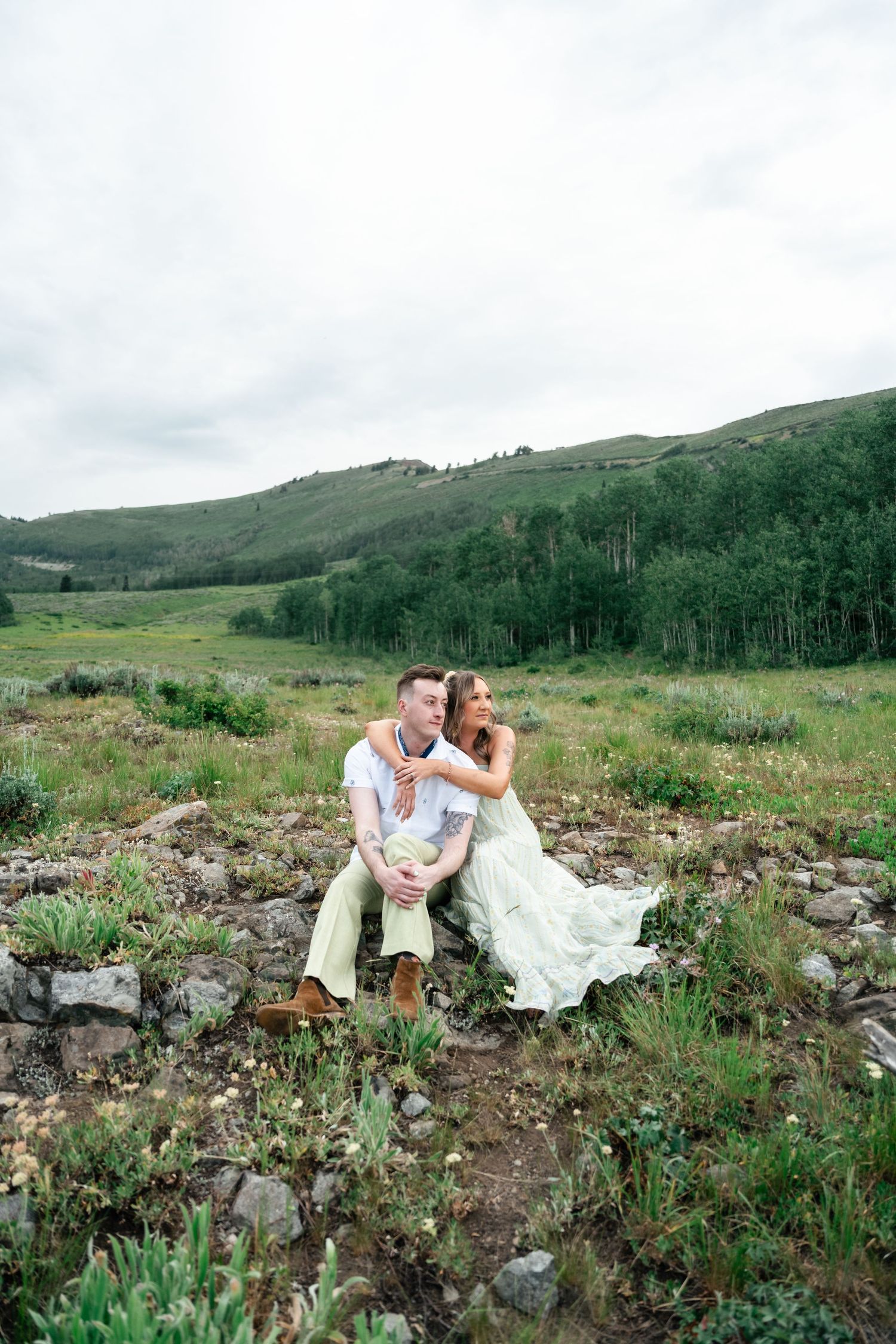 Couple in wedding attire sits together on rocky mountainside with aspen trees and green hills in background. Utah mountain engagement pics.