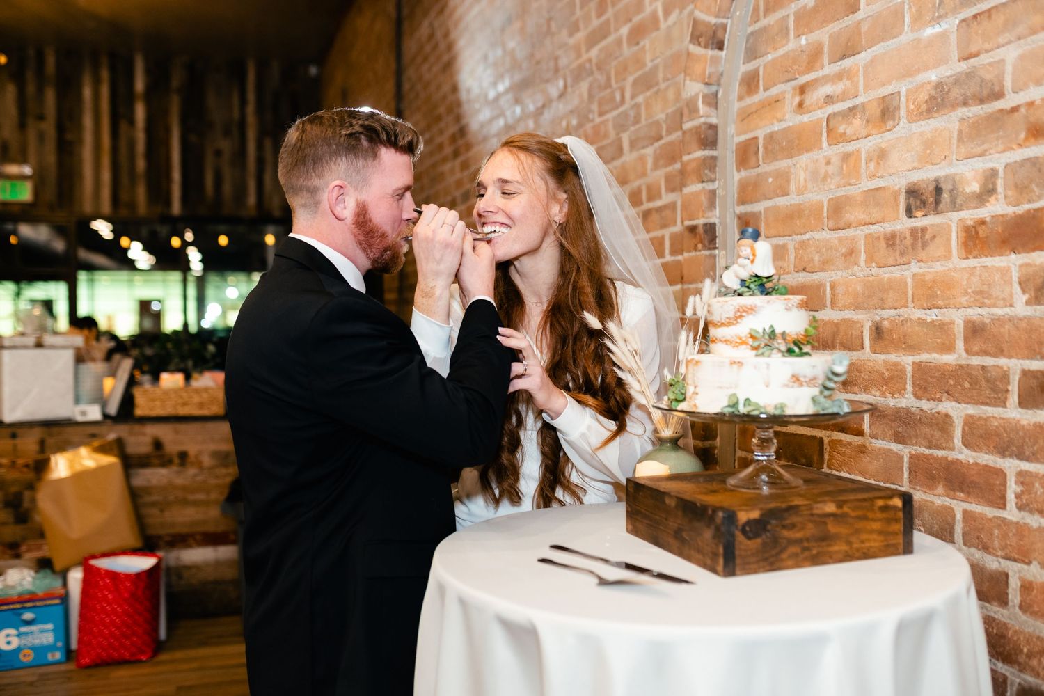 A couple shares a playful moment while cutting their wedding cake in front of an exposed brick wall.