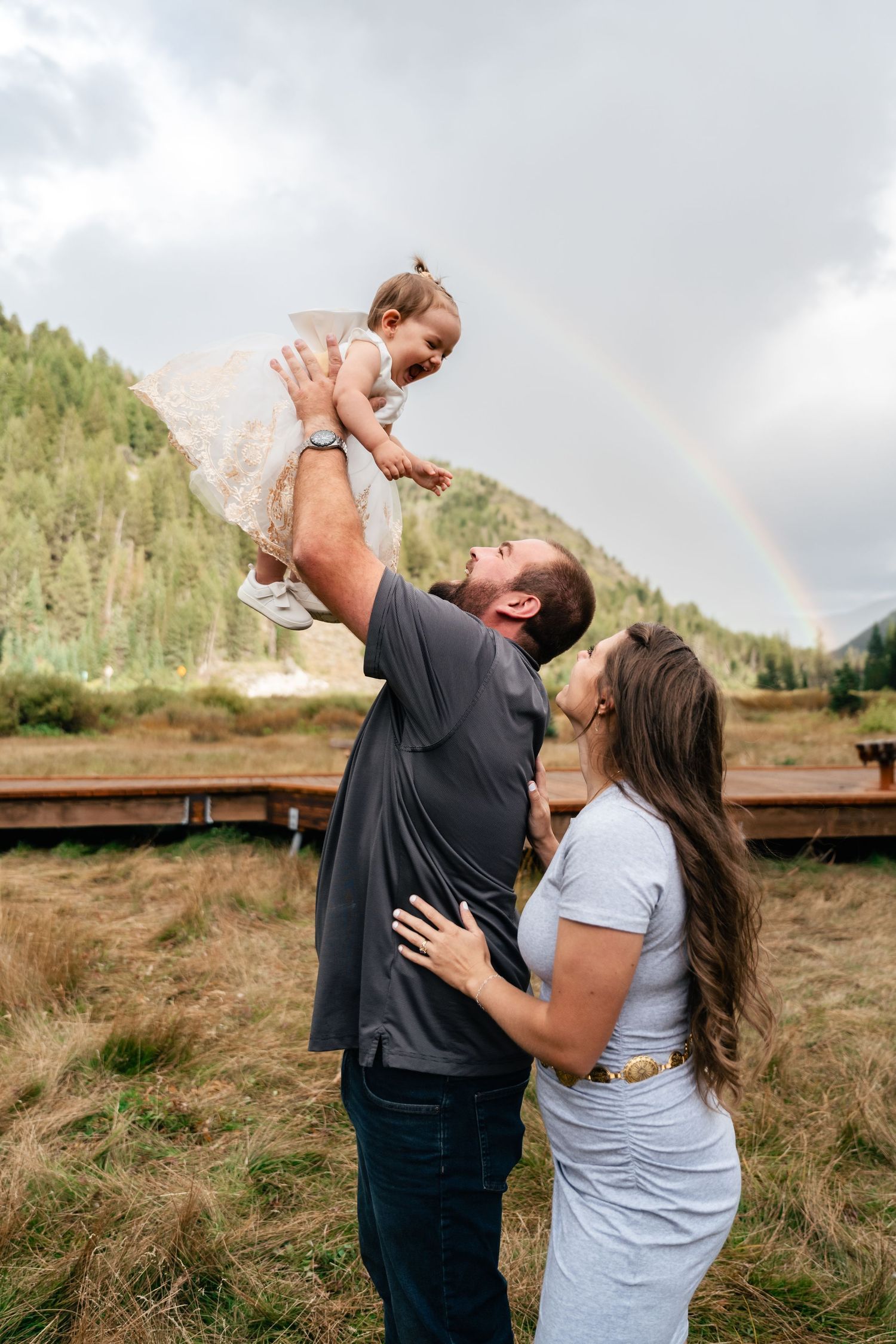 A family shares a tender moment with a rainbow in the background of a mountain landscape.