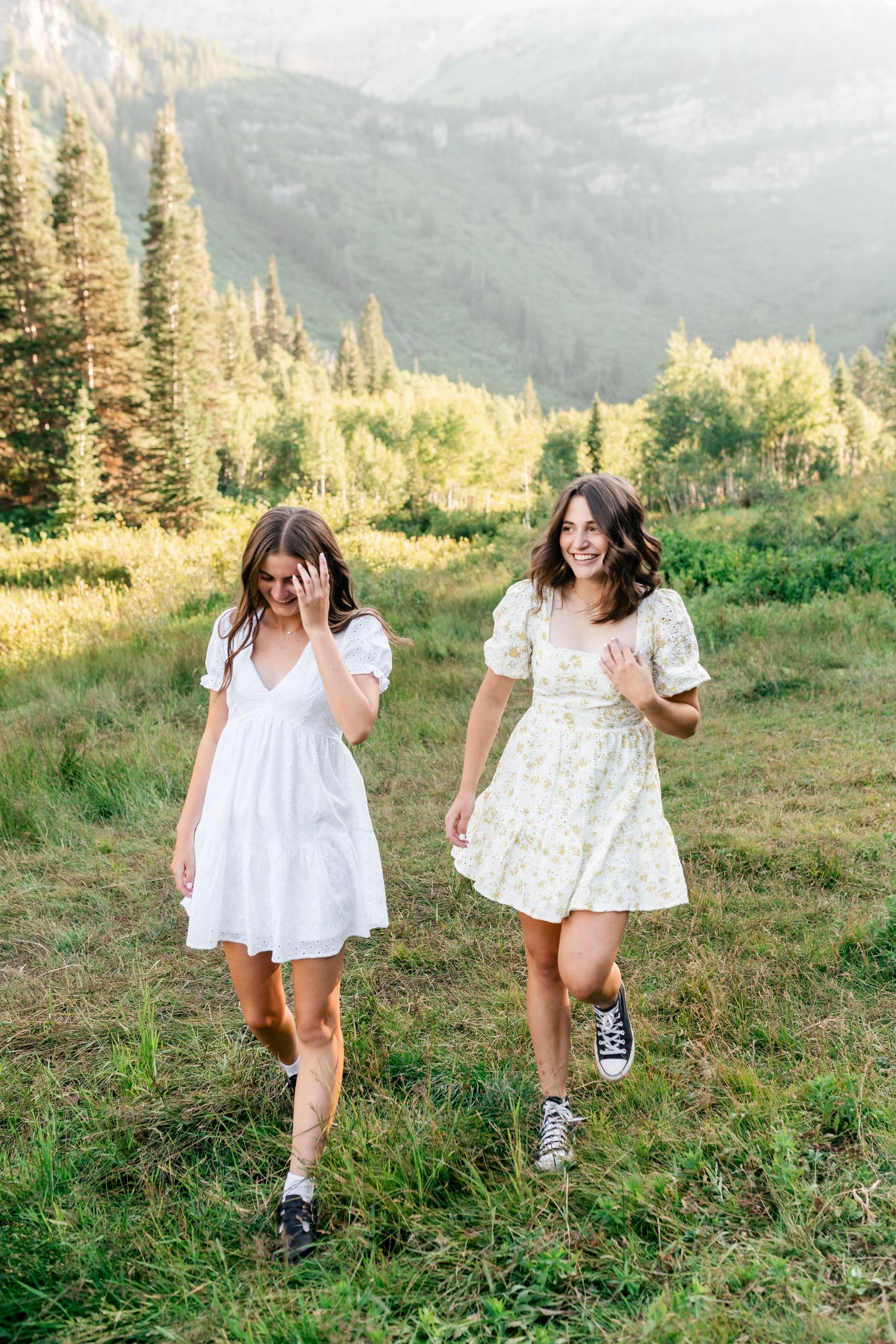Two people in white dresses and sneakers playfully run through a mountain meadow at sunset.