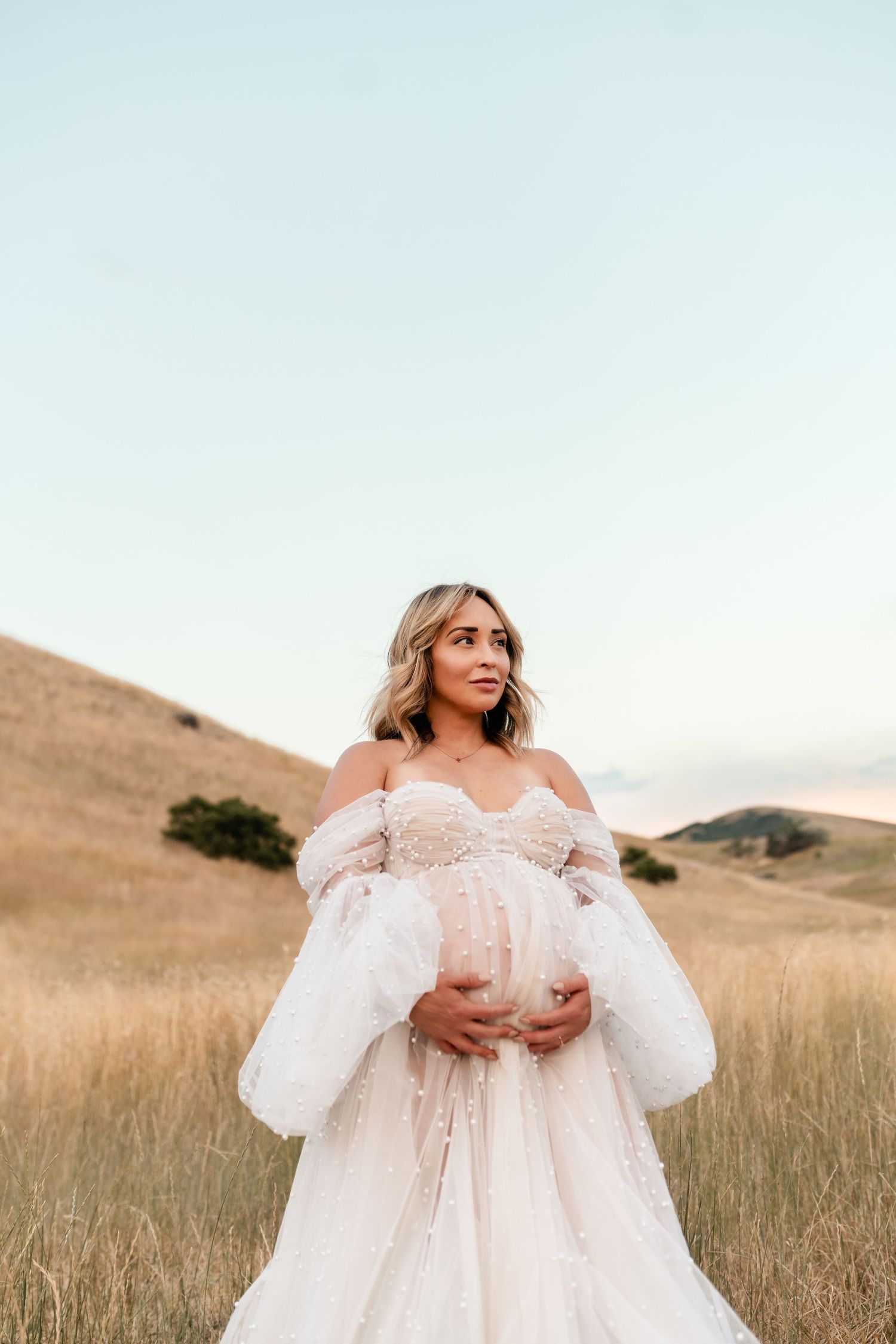 A pregnant woman in a flowing white lace gown poses in a golden field at sunset with rolling hills in the background.