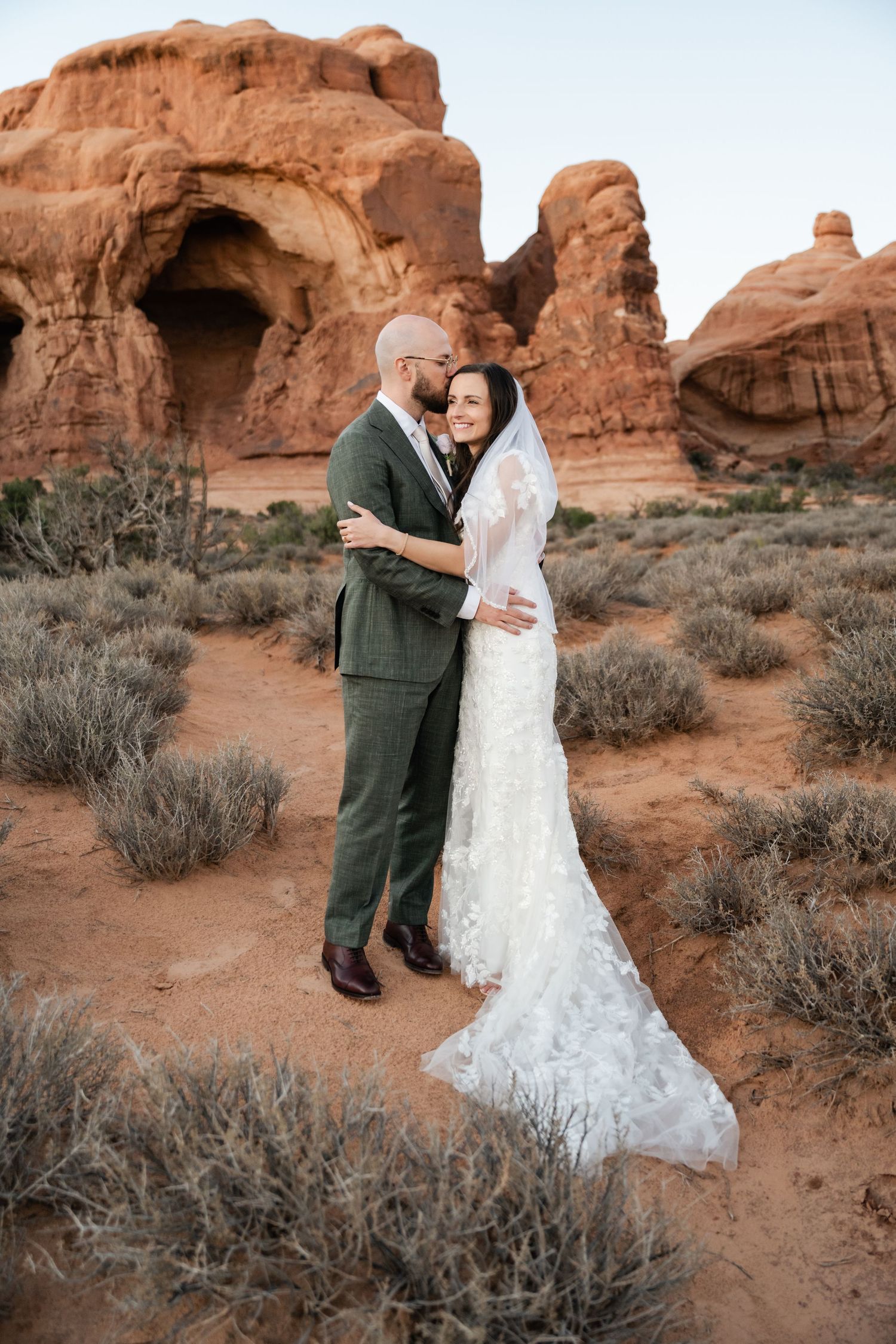 A couple shares an intimate moment among red rock formations and desert brush at sunset in Arches National Park.