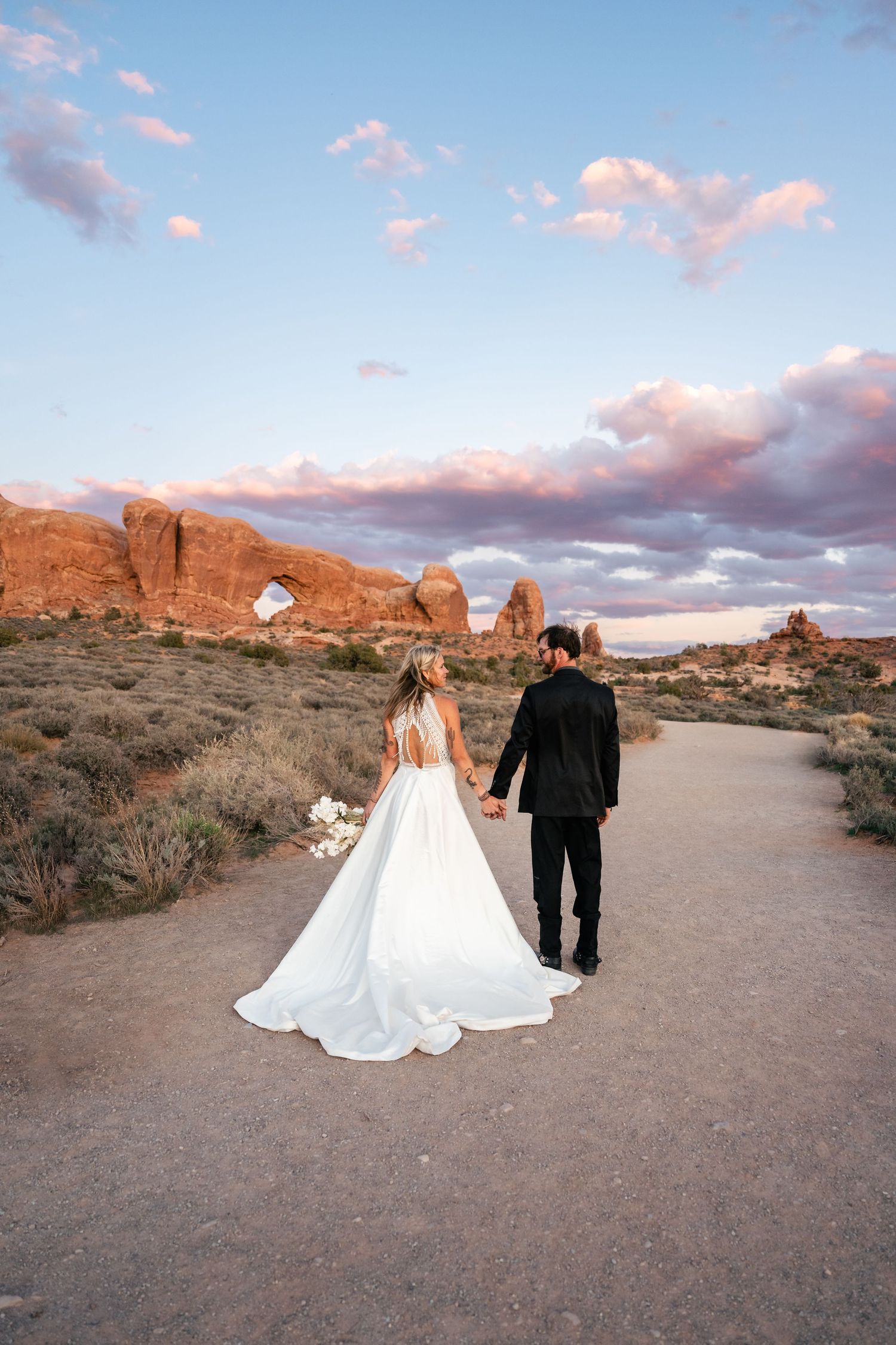 A couple walks along a desert path surrounded by red rock formations and desert brush under a cloudy sunset sky.