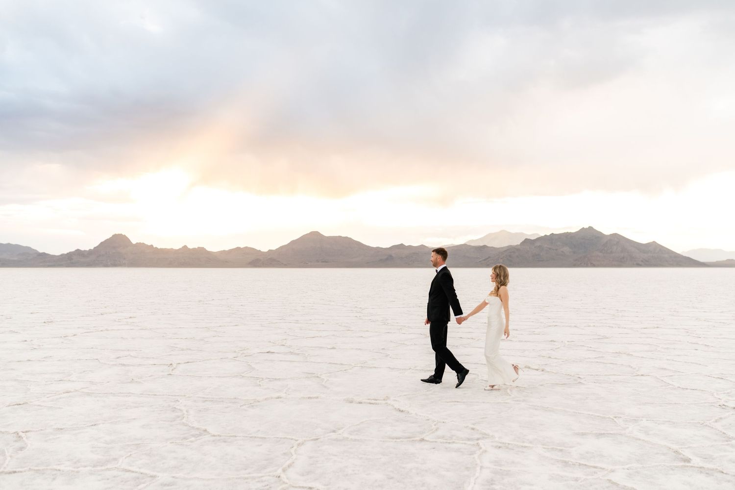 A couple walks hand in hand across the stark white Bonneville Salt Flats during sunset with mountains in the distance. Salt Flats Elopement.