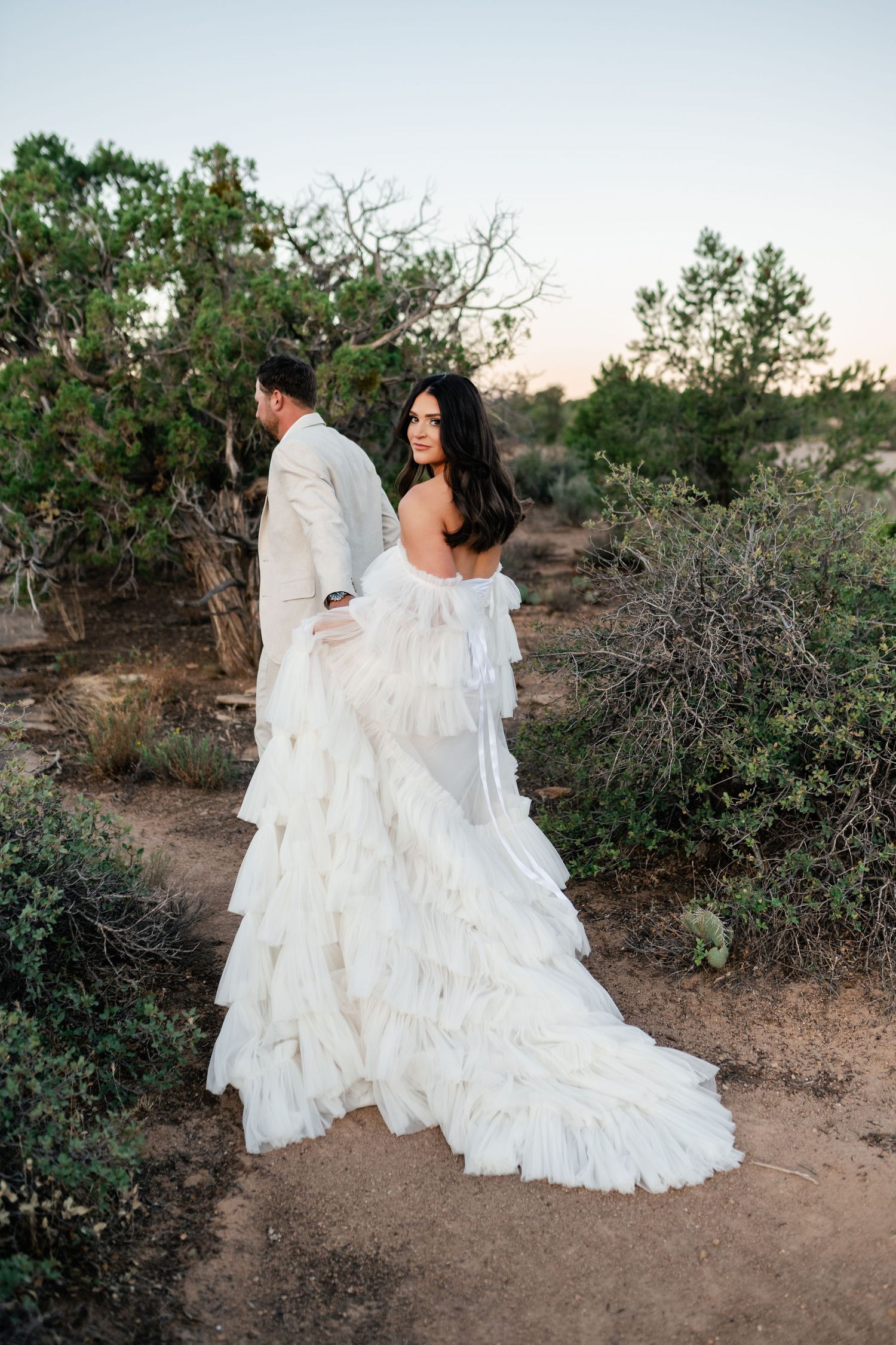 Zion Adventure Session. A flowing white wedding dress trails behind a couple as they embrace in a desert landscape with shrubs.