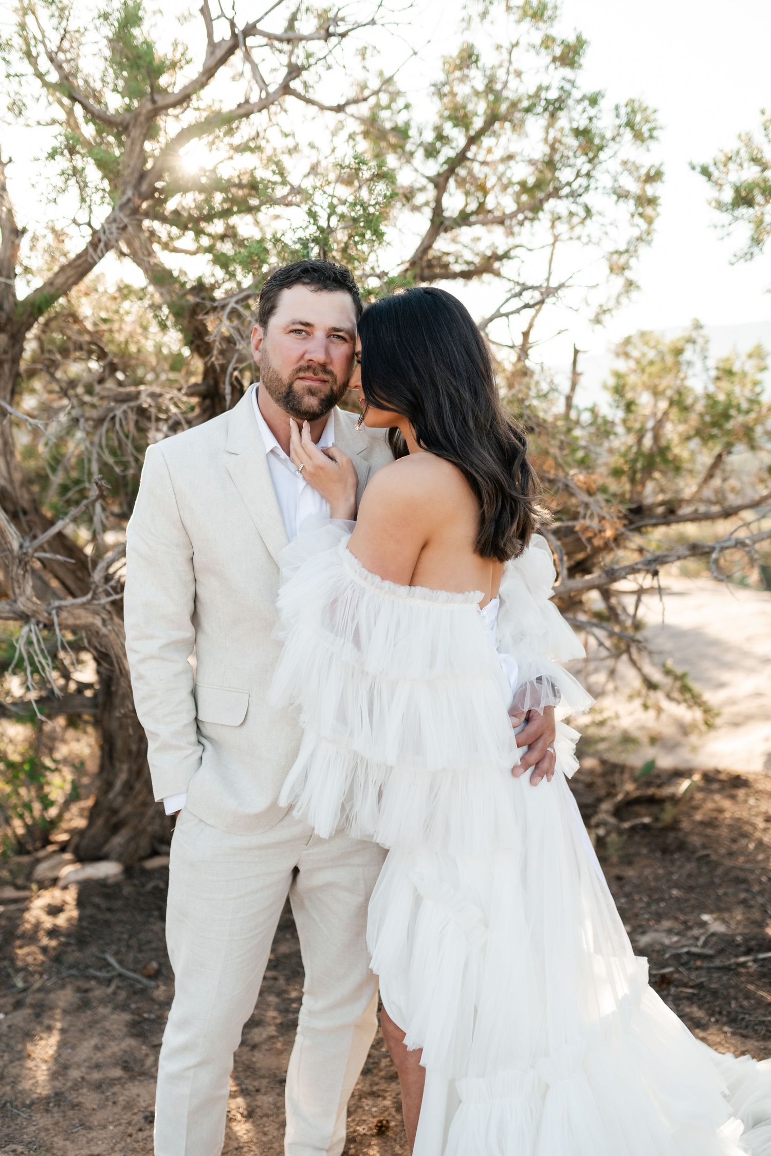 Zion destination photos. A couple shares an intimate moment outdoors wearing elegant white wedding attire against a natural desert backdrop.