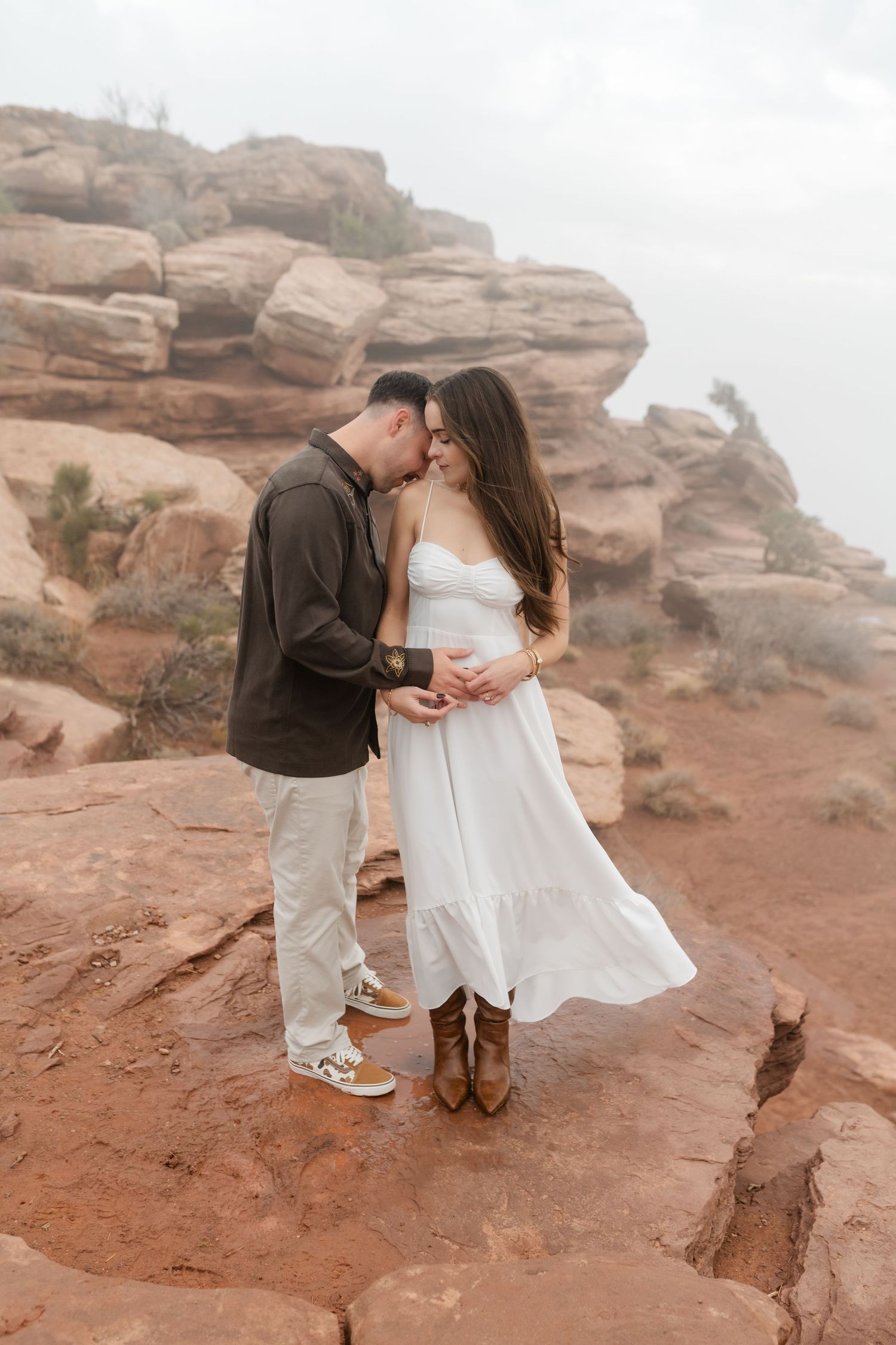 A romantic couple embraces on red rock formations in a desert landscape during sunset.