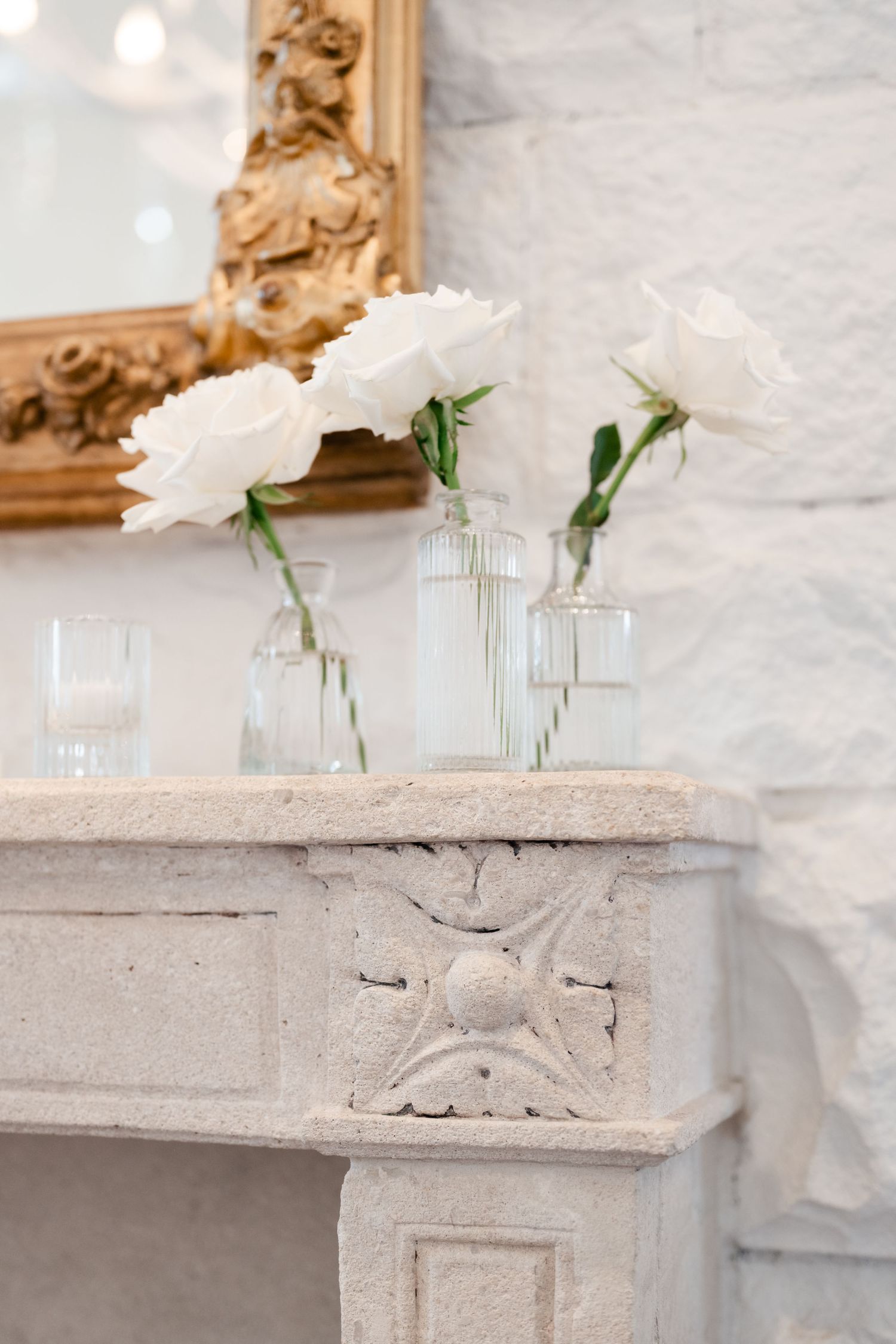 White peonies in clear glass vases displayed on an ornate stone mantel with gold-framed mirror.