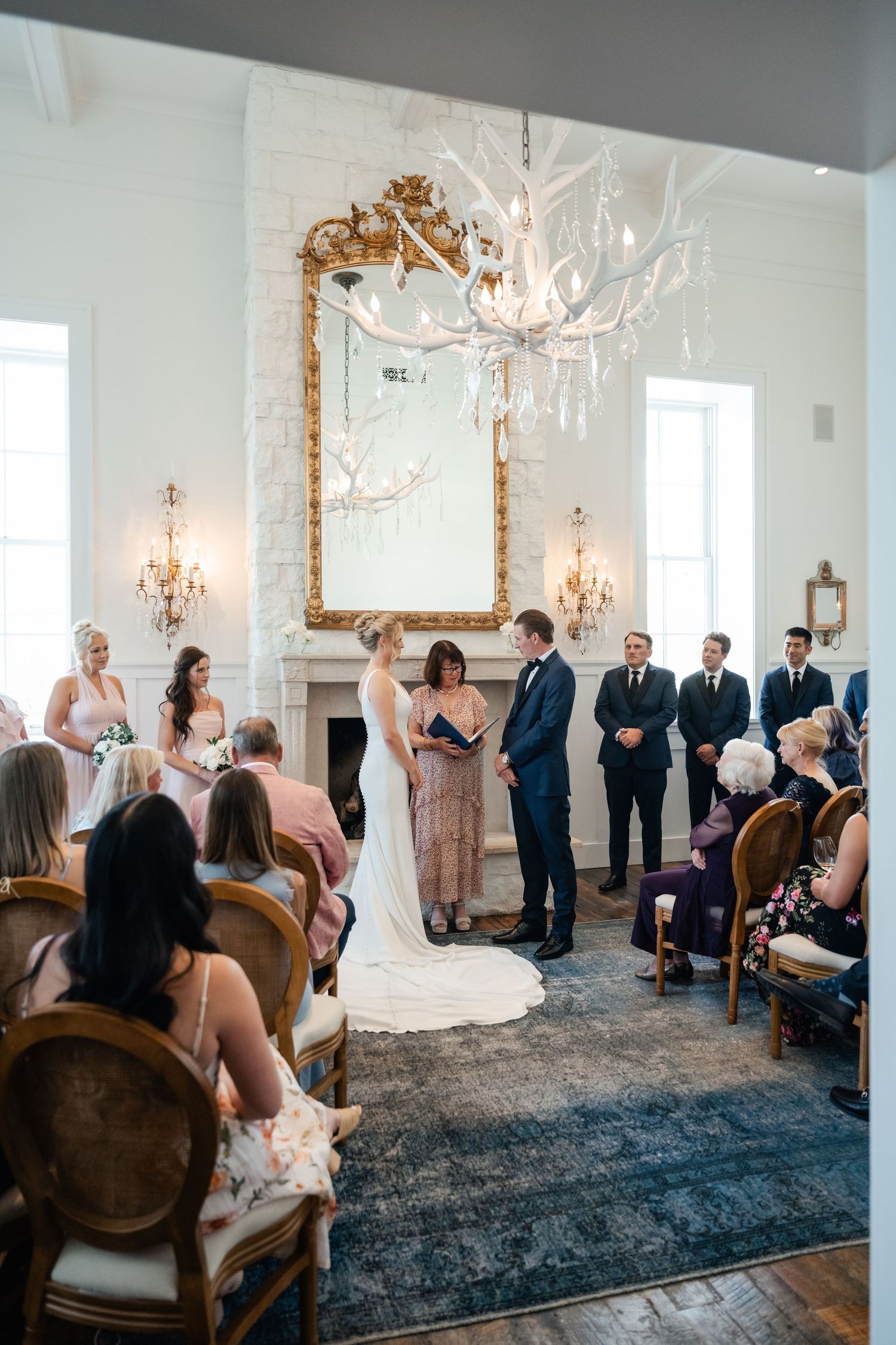 Wedding ceremony taking place in an elegant white room with chandeliers and a large ornate gold mirror.