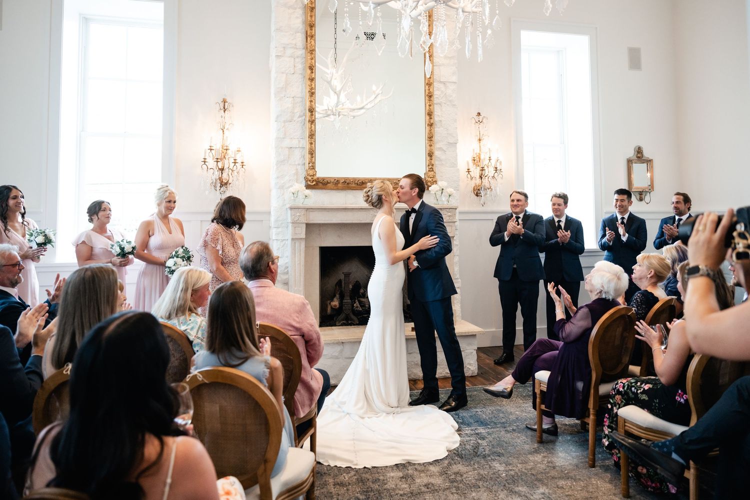 Wide view of wedding ceremony in bright white room with guests seated on both sides.
