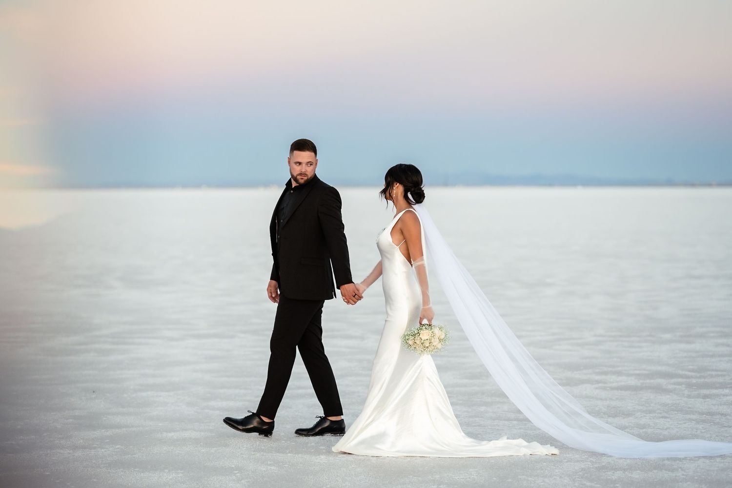 Couple in wedding attire creates artistic poses on salt flats at dusk.