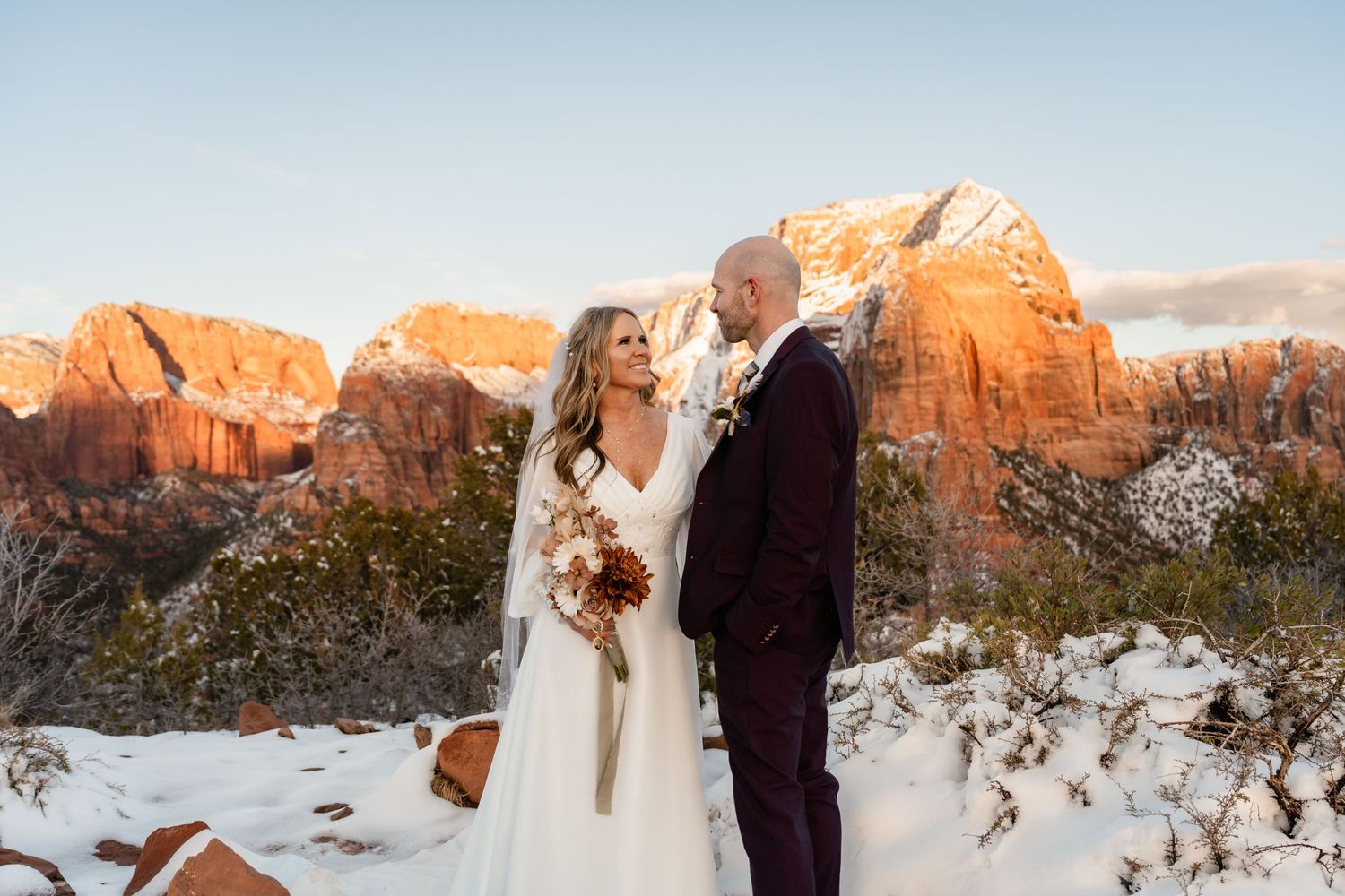 A wedding couple poses against orange cliffs and snowy terrain during golden hour.
