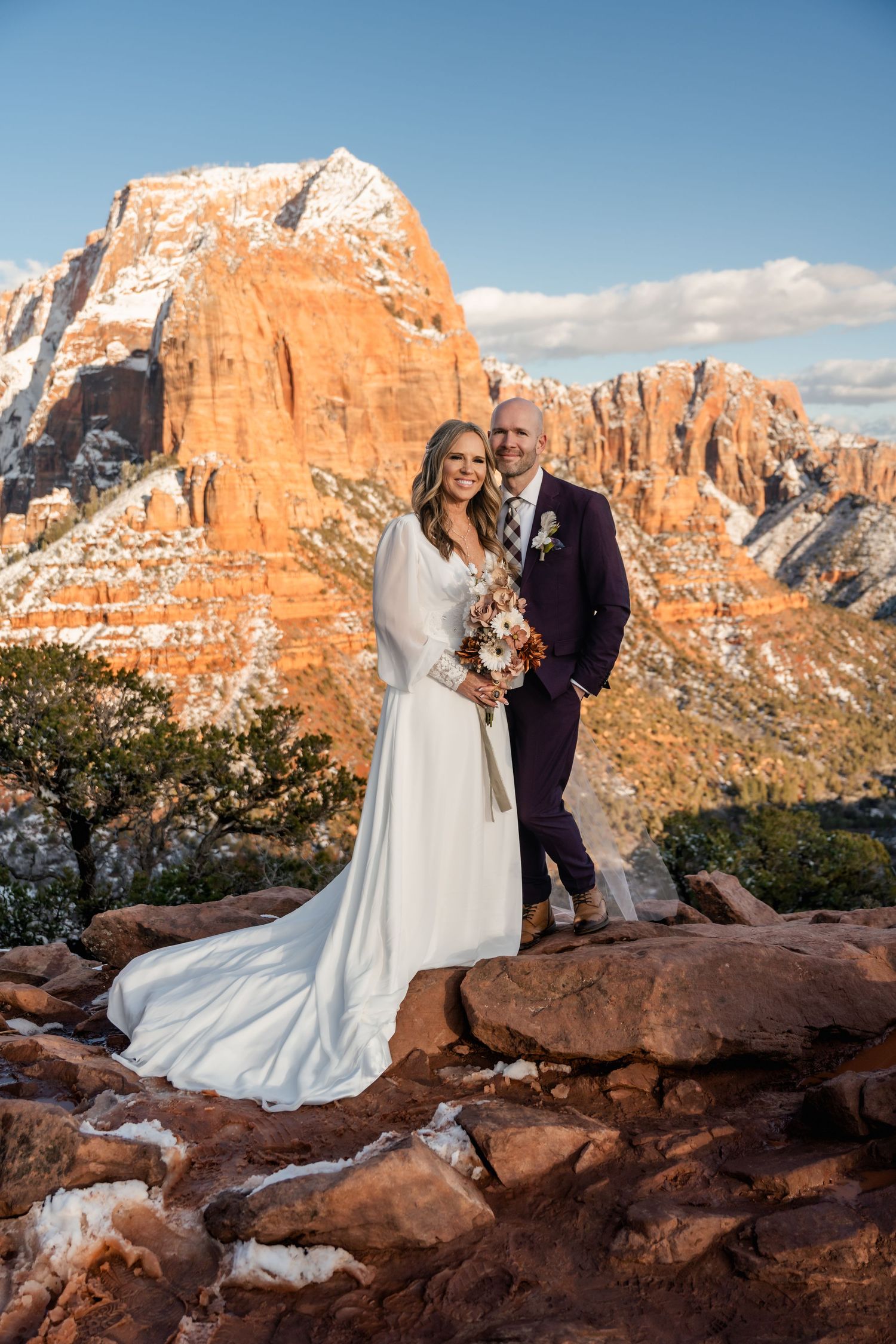 Wedding portrait against dramatic red rock cliffs and snow-capped mountains.