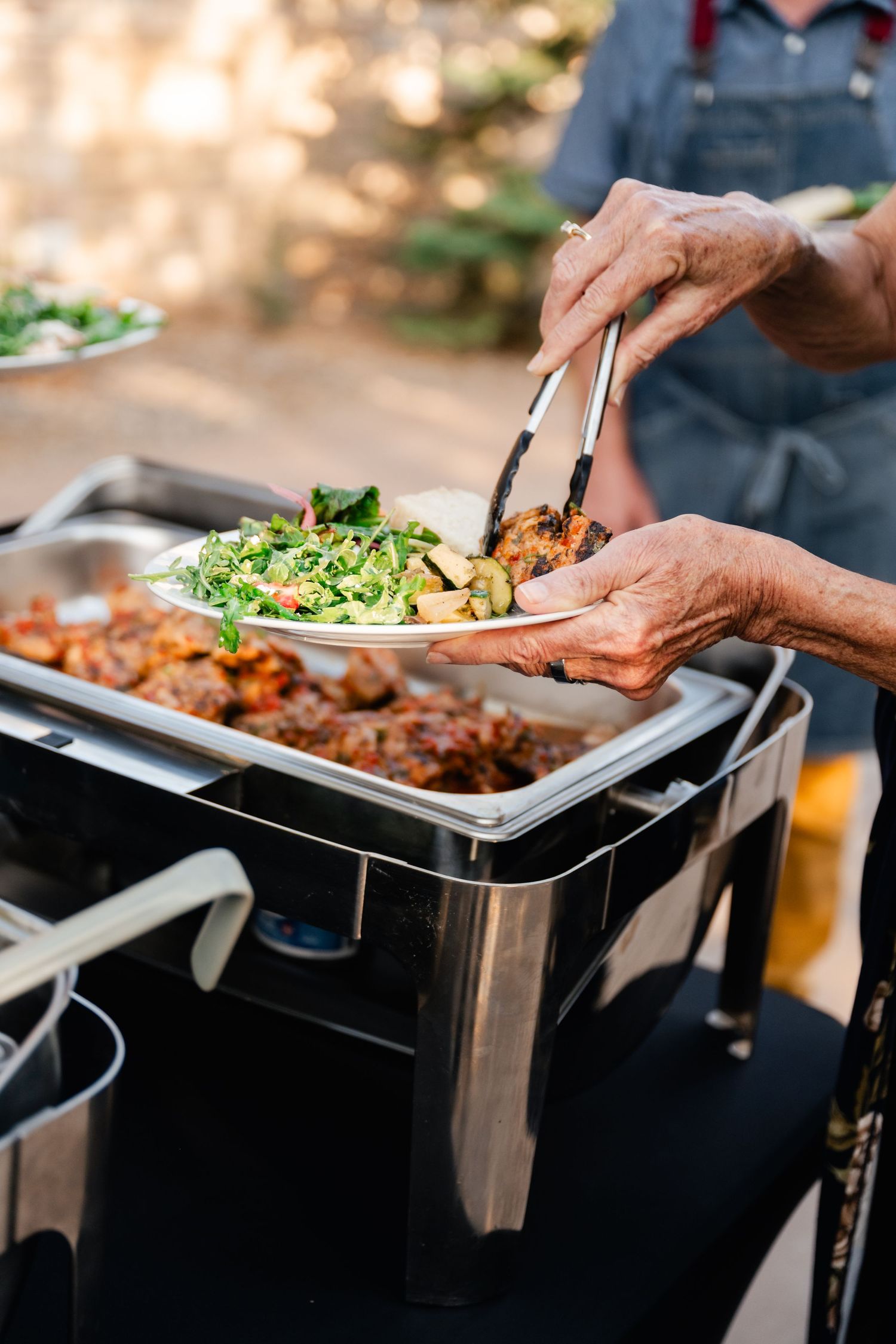 Catering staff serving food from stainless steel chafing dishes at an outdoor event.