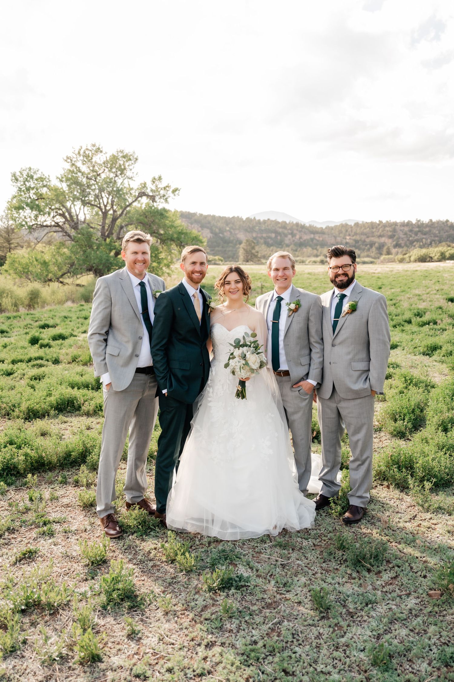 Wedding party poses in a scenic field with rolling hills and trees in the background on a cloudy day.