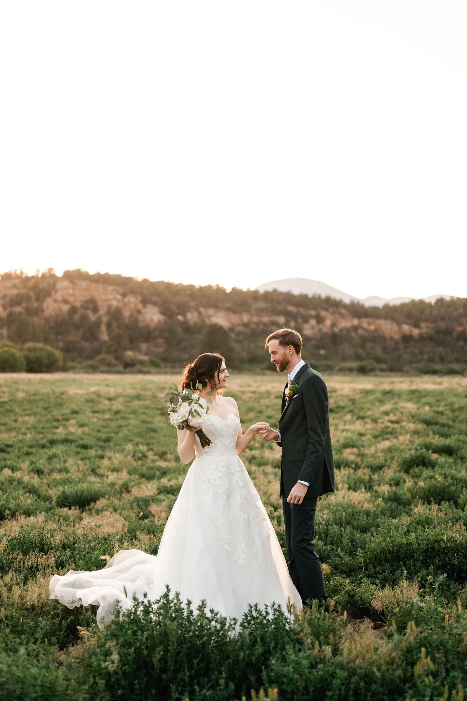 Newlyweds share an intimate moment in a green meadow with mountains in the distance.