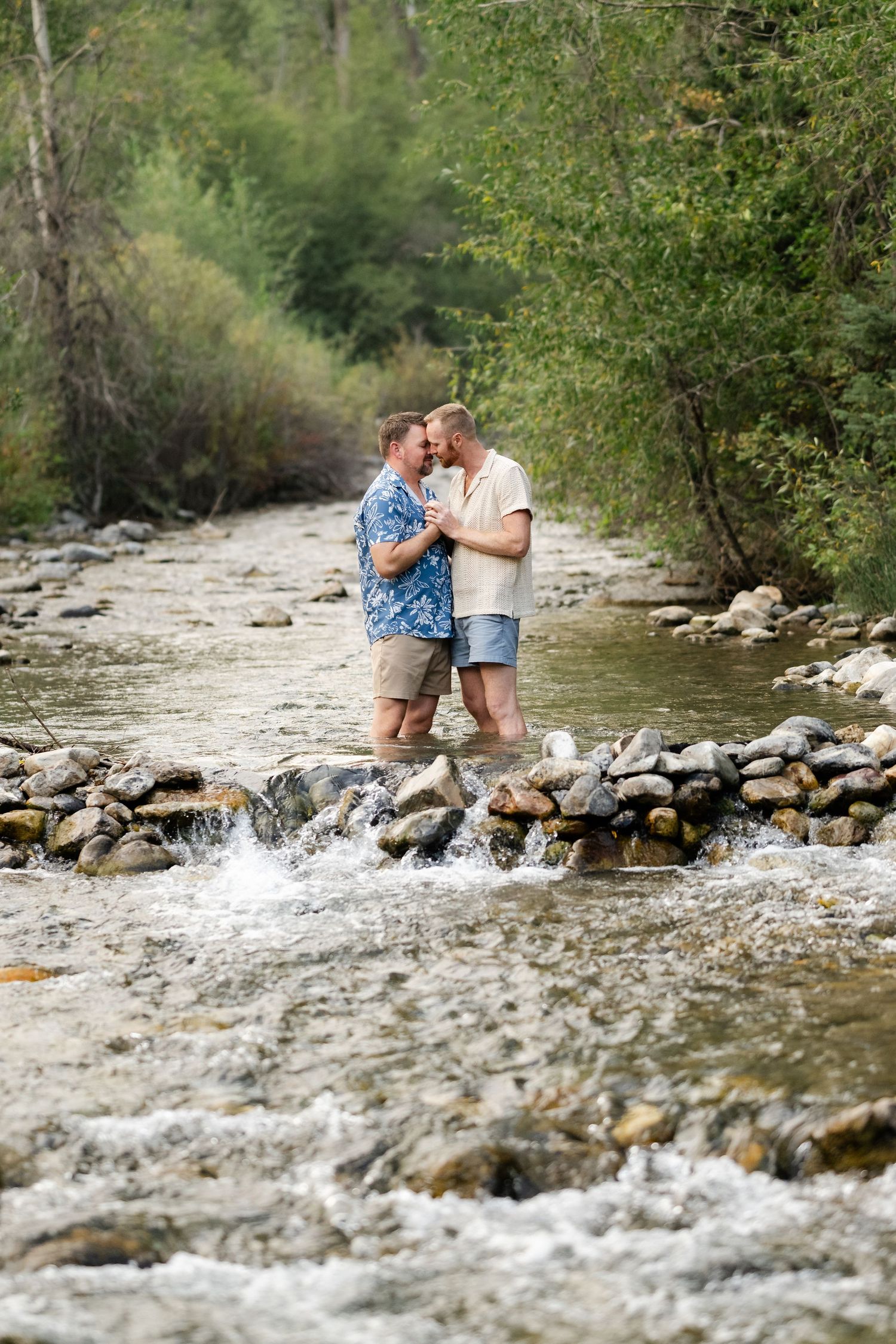 Two men sharing a romantic moment standing on rocks in a flowing mountain stream surrounded by lush greenery.