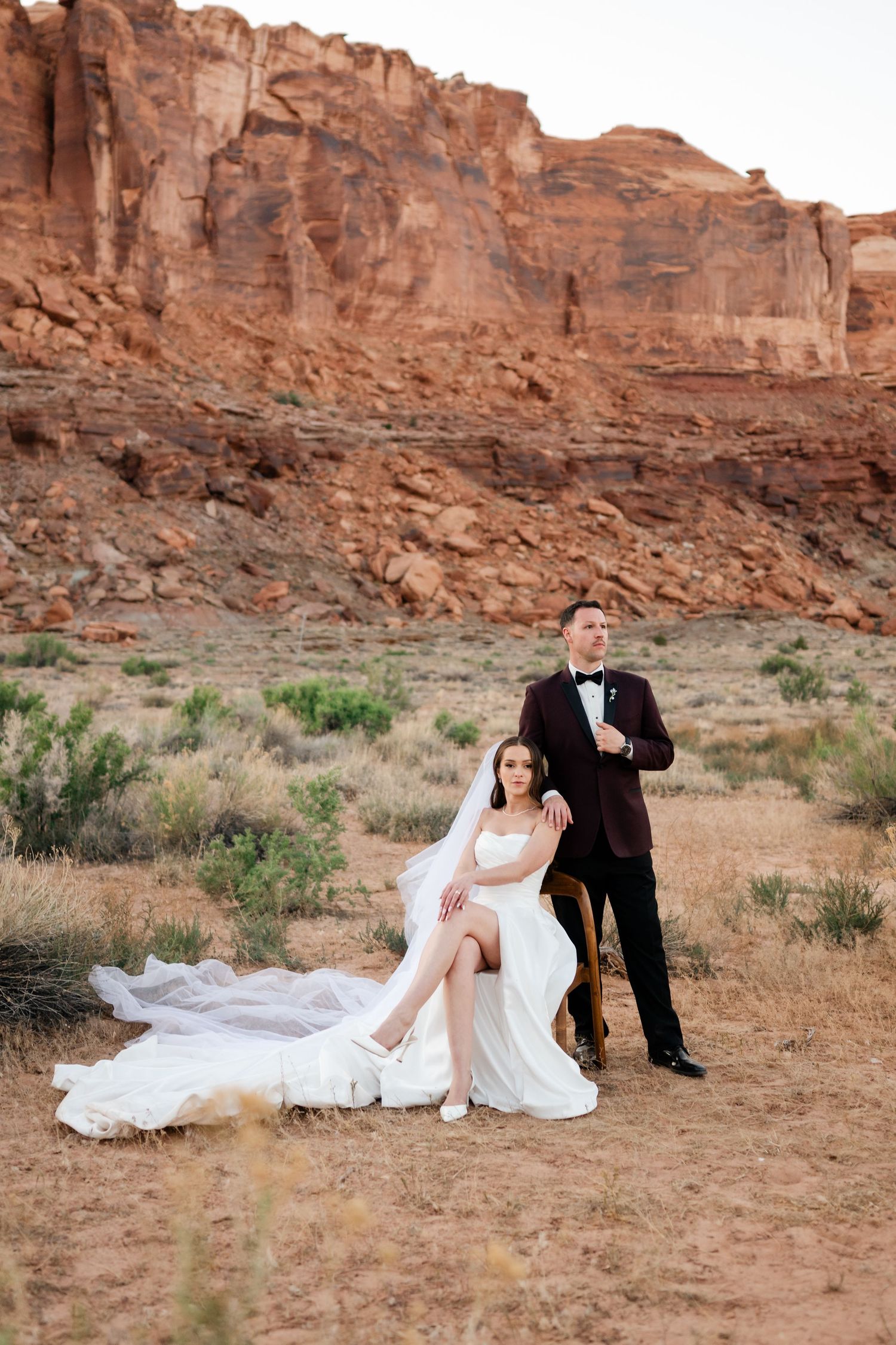 Bride in white gown sits while groom in burgundy tuxedo stands beside her against red rock canyon backdrop.