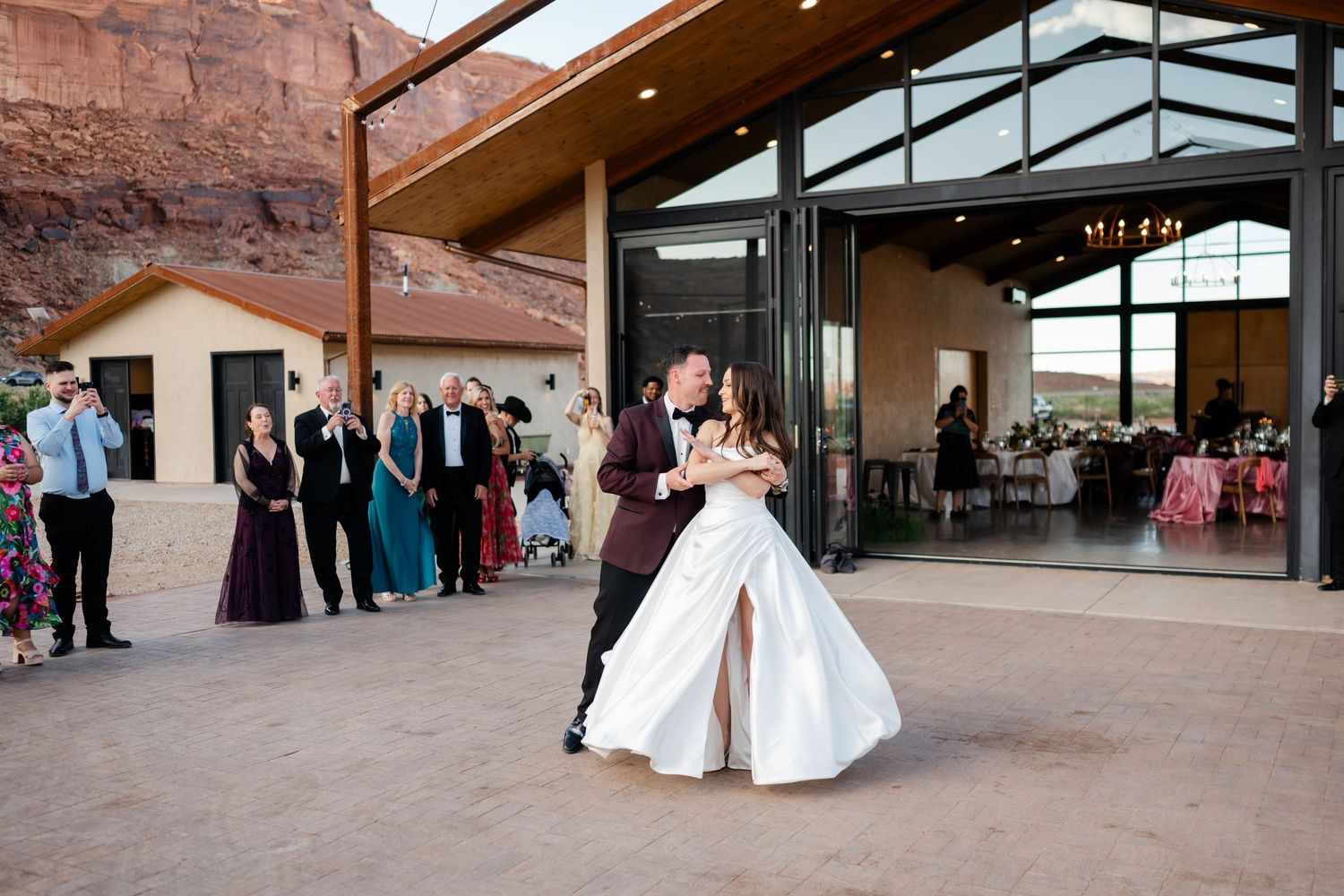Bride and groom share first dance outdoors at desert wedding venue with red rock canyon backdrop and guests watching.