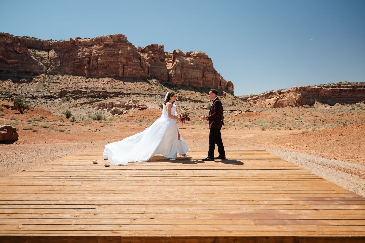 Bride and groom exchange vows on wooden deck amid red rock desert landscape under clear blue sky.