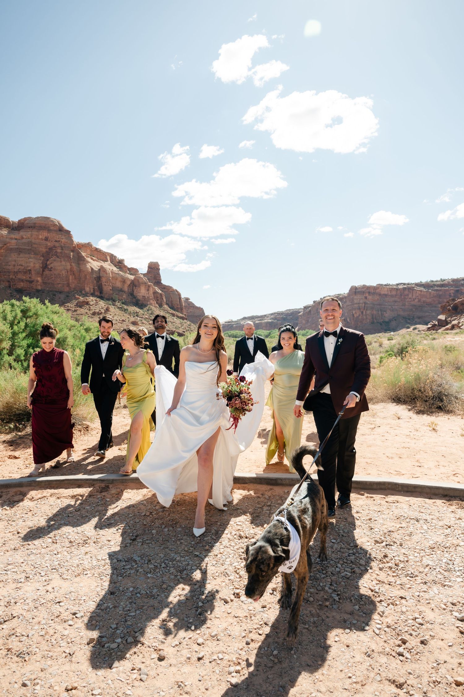Wedding party walks through red rock desert landscape, bride in white gown holding bouquet, groom in tuxedo leading a dog.