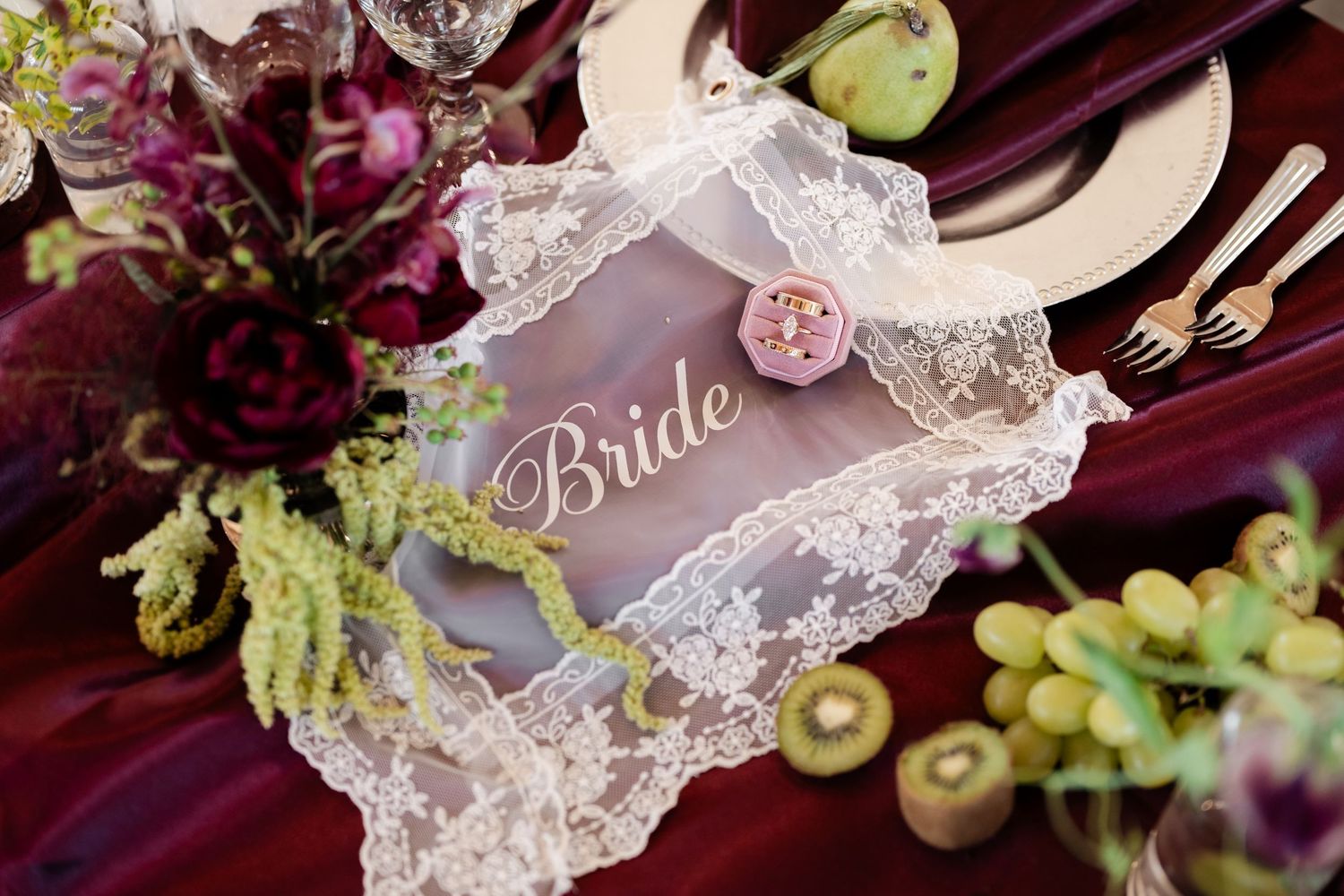 Elegant bride place card with lace trim, pink rose, and gold lettering on a burgundy wedding table with fruit accents.