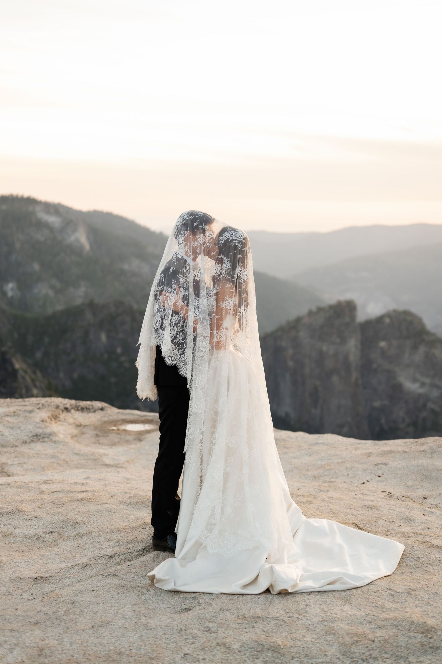 Bride and groom share a kiss under a lace veil on a rocky mountain overlook at sunset.