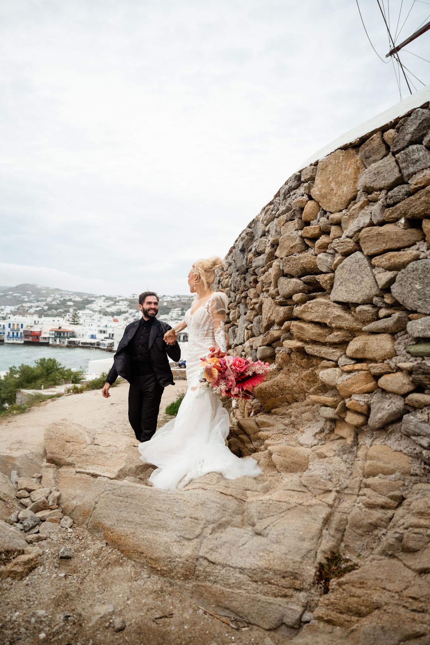 Bride and groom walk along a stone path in Mykonos, Greece, with whitewashed buildings and sea views behind them.