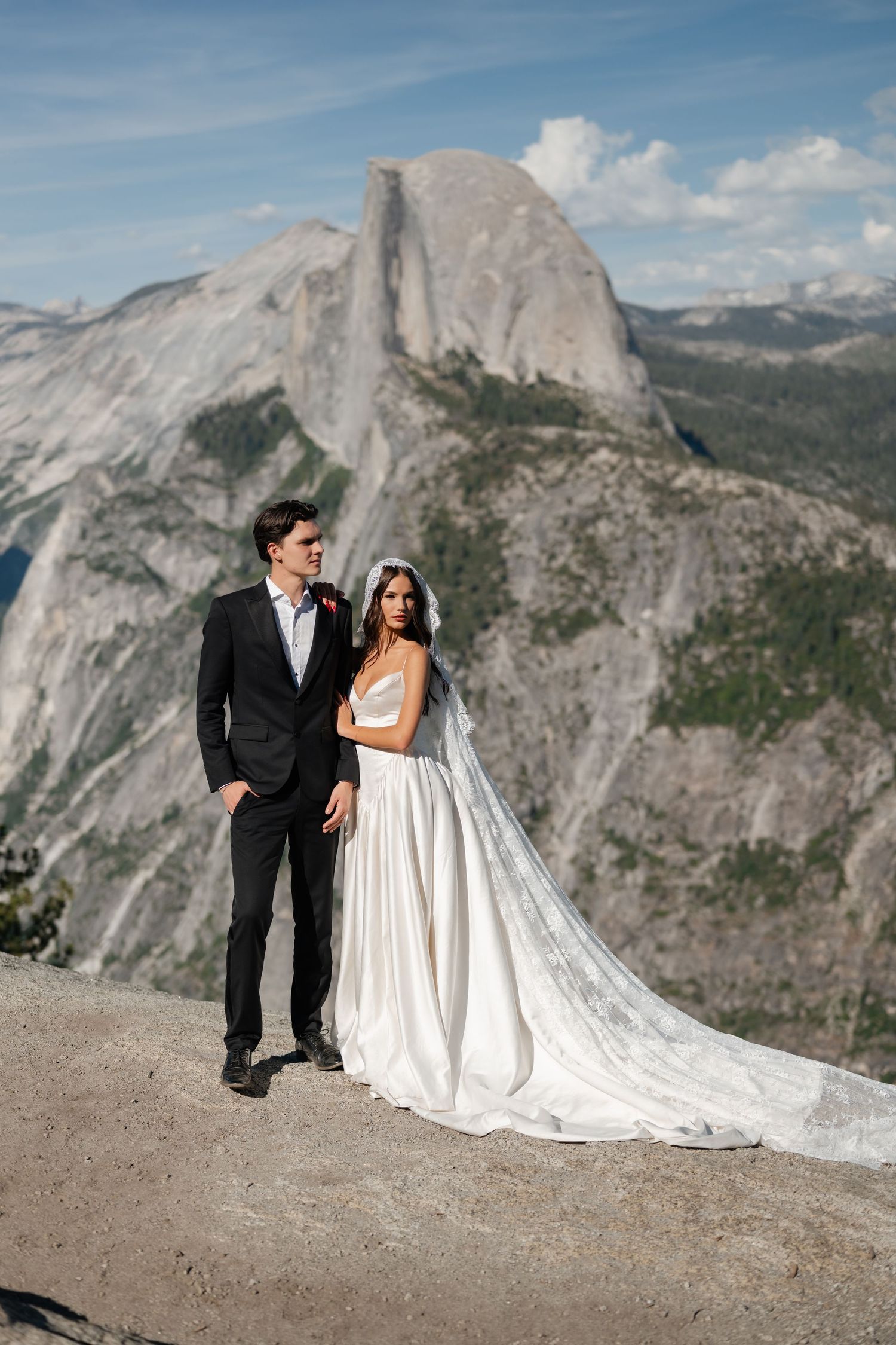 Bride and groom pose on rocky overlook with Half Dome at Yosemite National Park behind them on their wedding day.