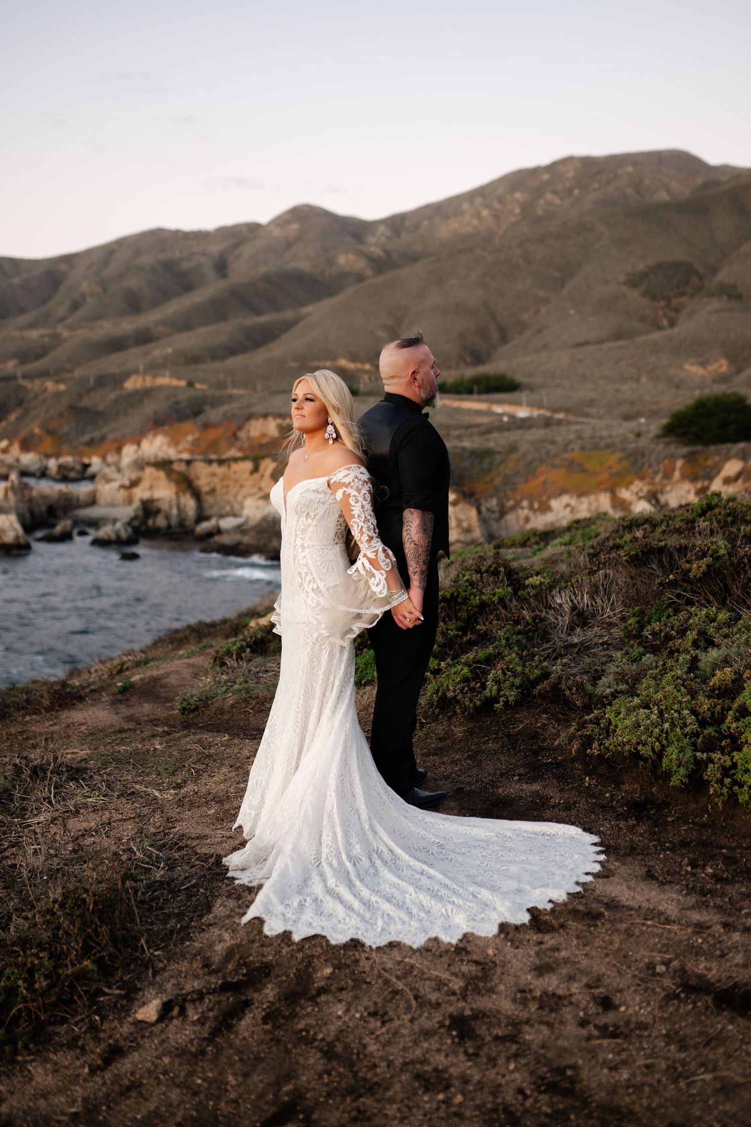 A couple in wedding attire poses against a scenic coastal mountain backdrop at sunset.
