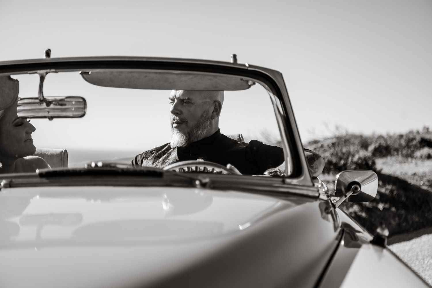 Black and white photo of classic car driver against coastal landscape.