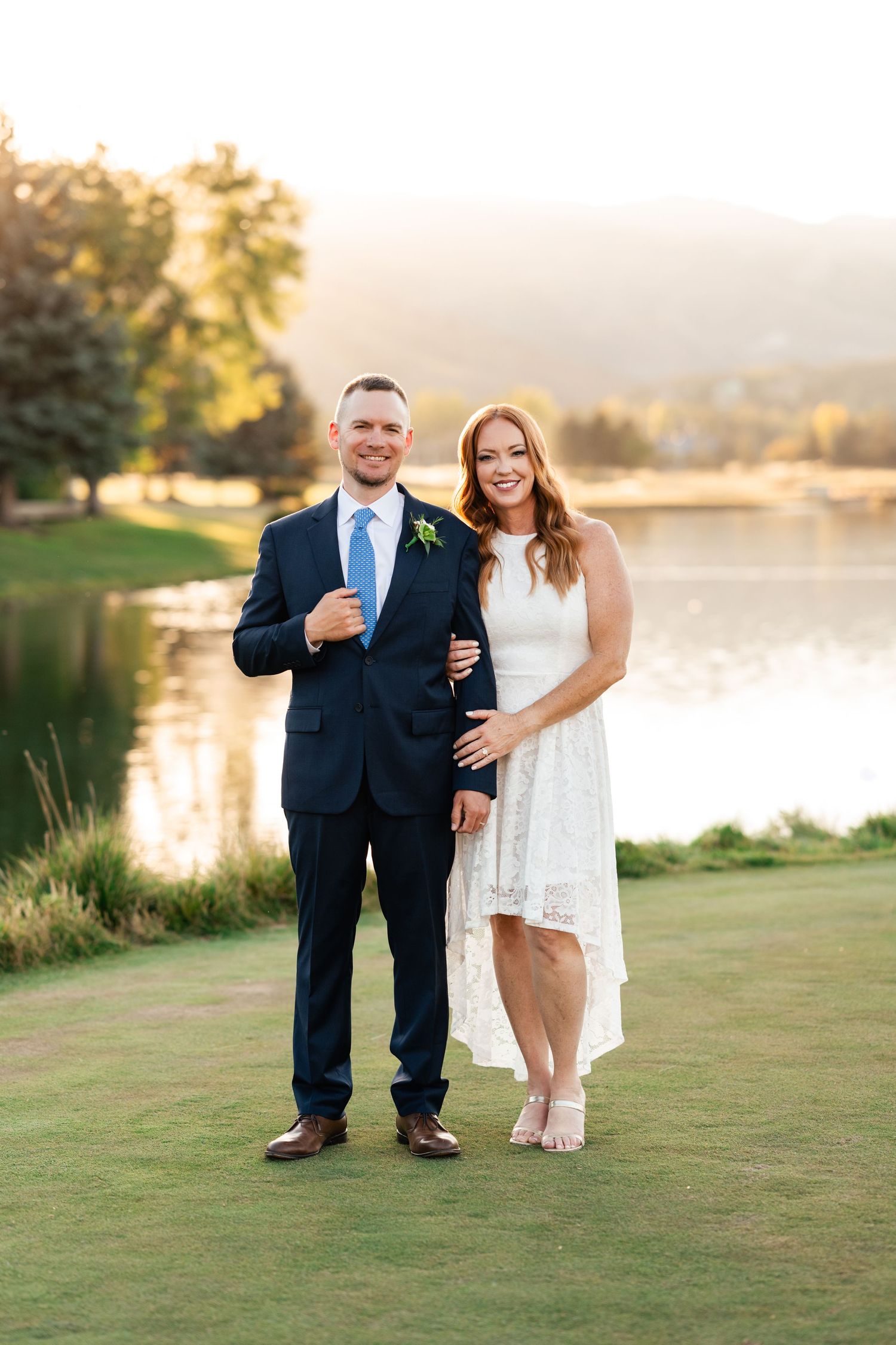Wedding couple poses on grassy lawn beside serene lake at sunset.