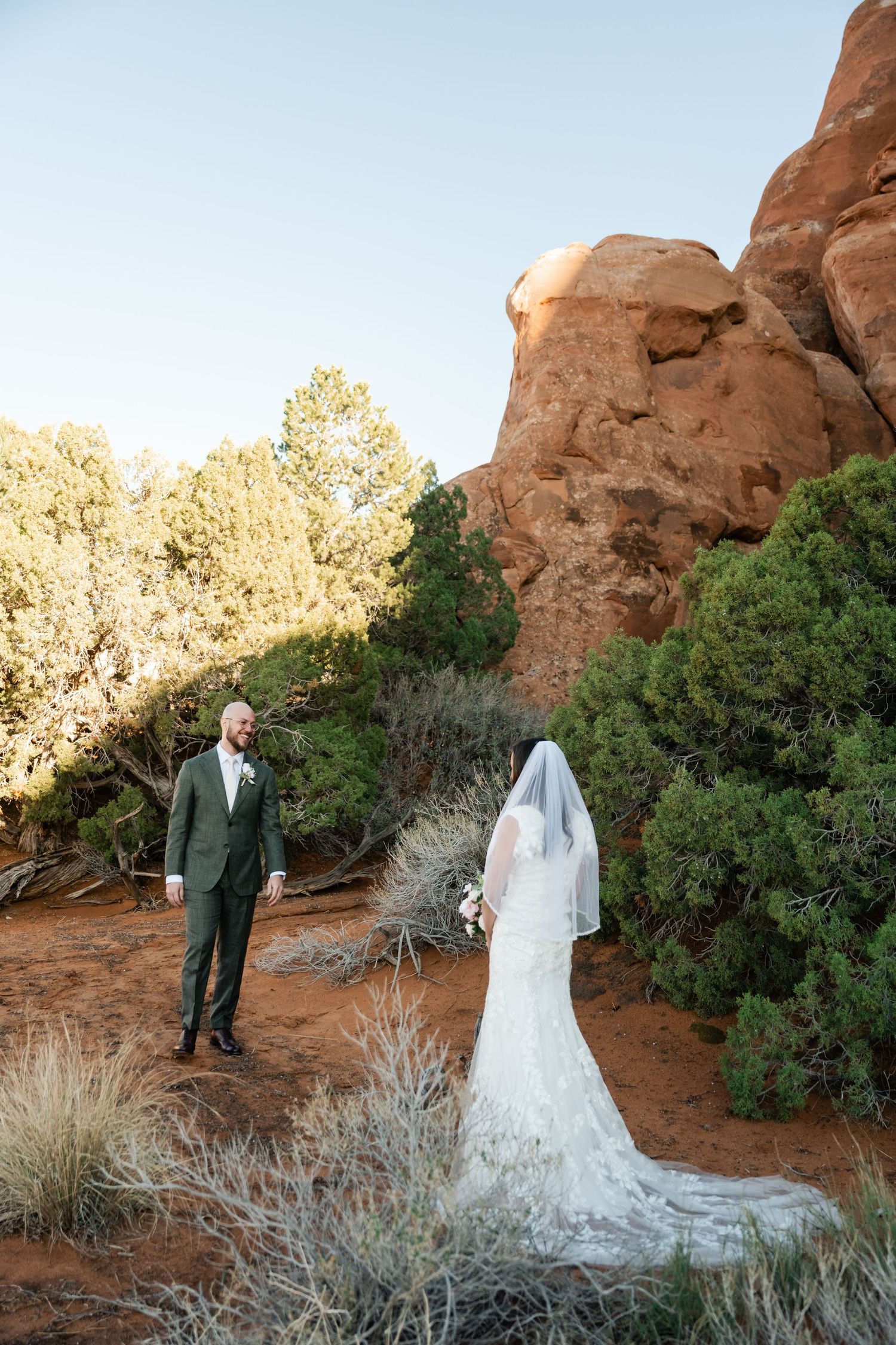A wedding couple stands among desert brush with red rock formations in the background.