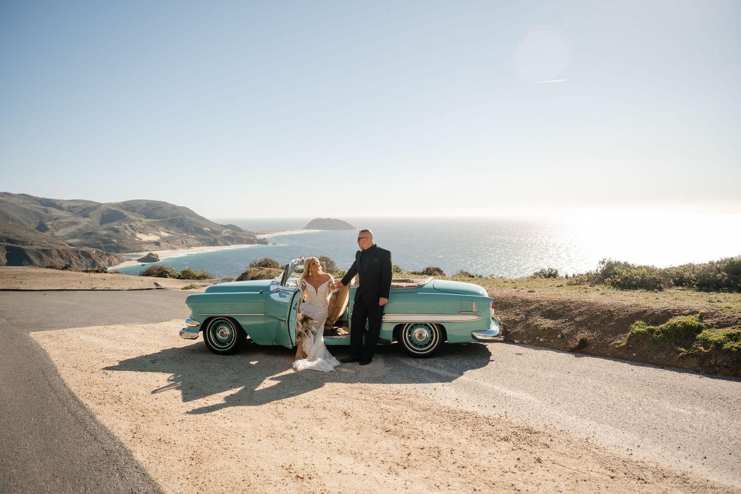 A vintage turquoise car parked along a scenic coastal road with mountains and ocean views in the background.