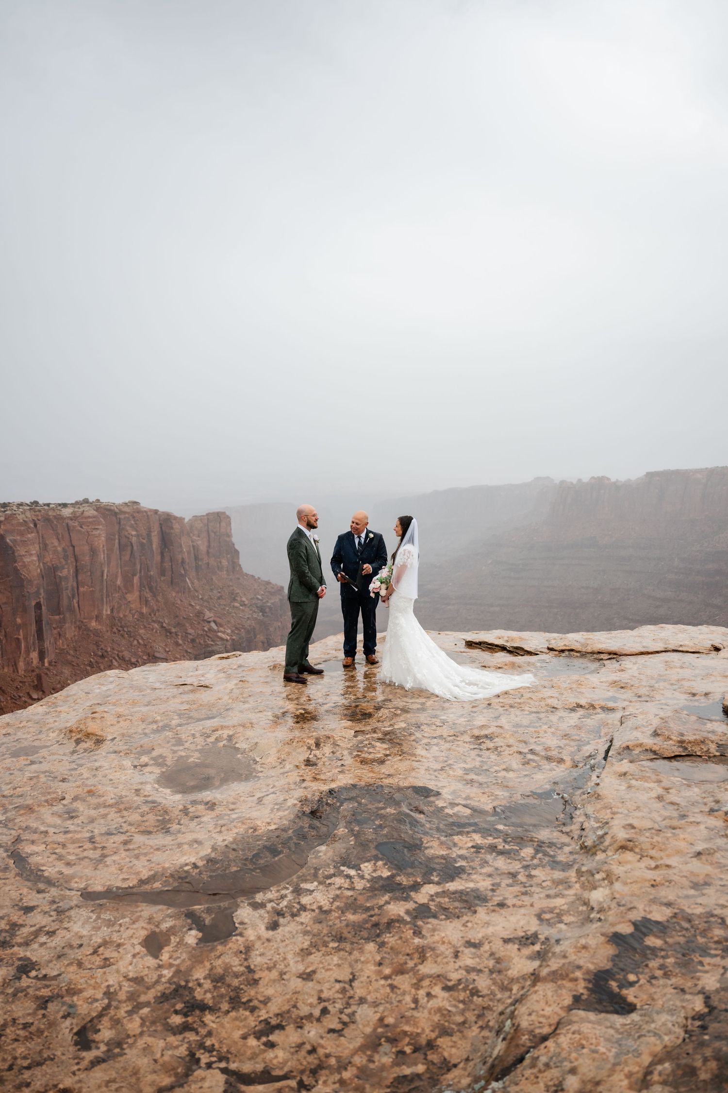 A wedding ceremony takes place on a dramatic cliff edge overlooking a misty desert canyon landscape.