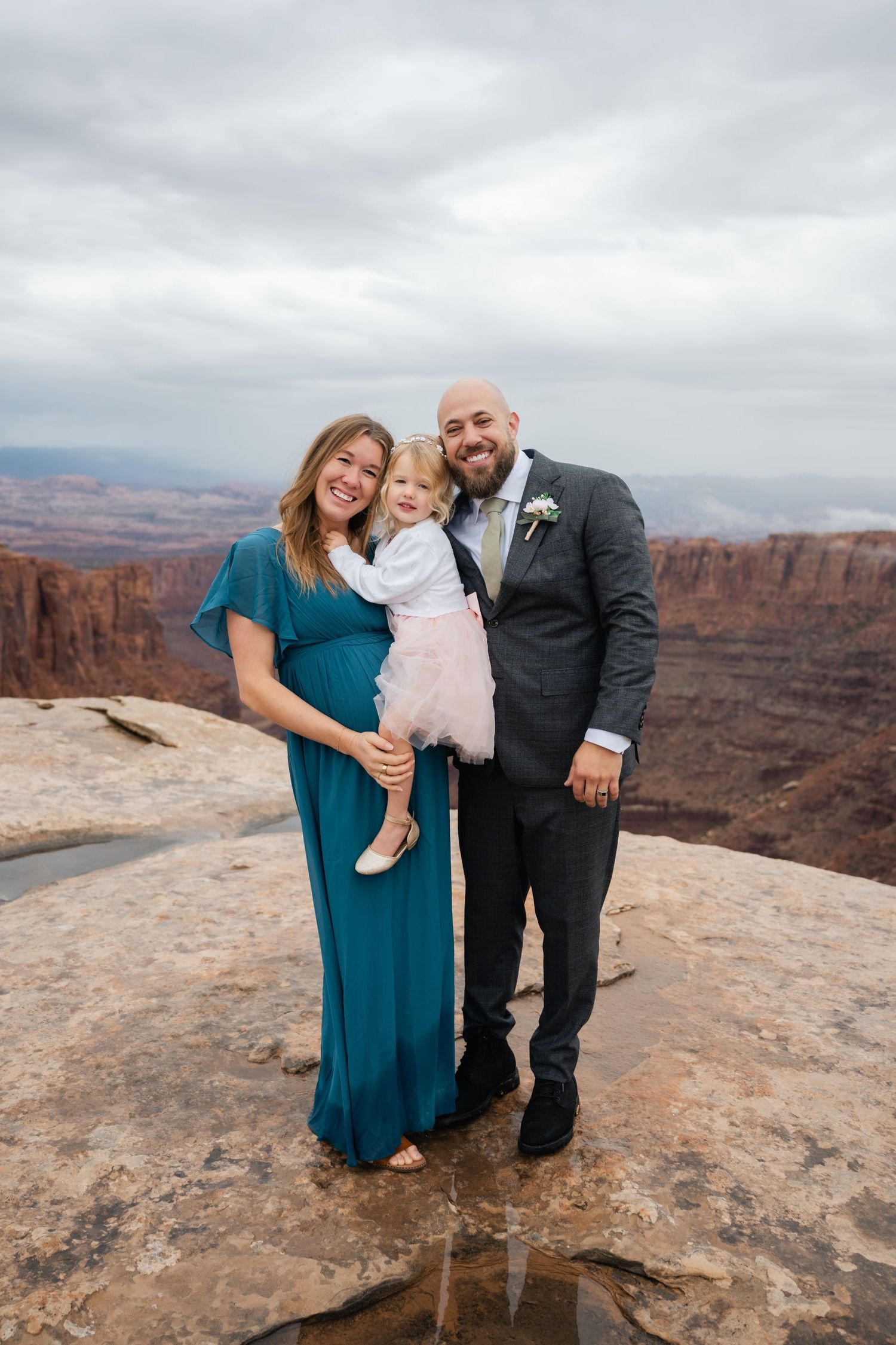 A couple and their white dog pose for photos against the dramatic backdrop of red rock canyons on an overcast day.