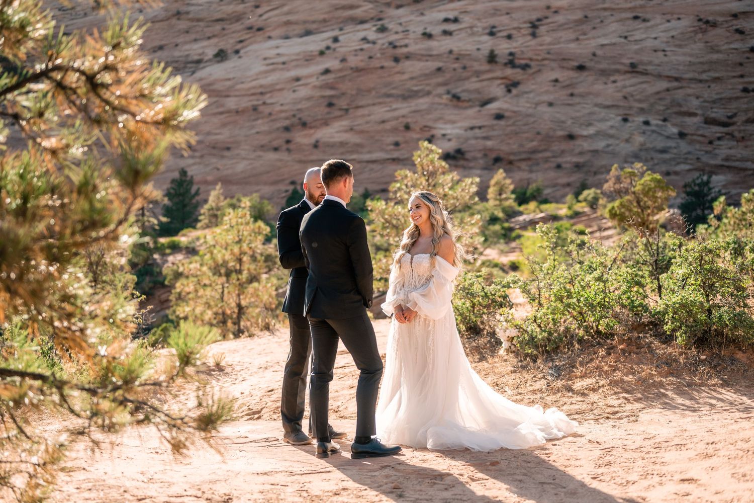 A couple in wedding attire stands on a scenic trail surrounded by desert vegetation.