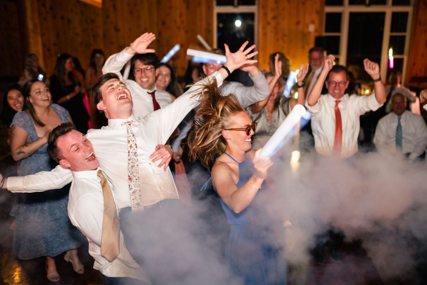 Party guests dance and celebrate at a wedding reception as smoke fills the dance floor beneath dramatic lighting. Utah Wedding Photographer.