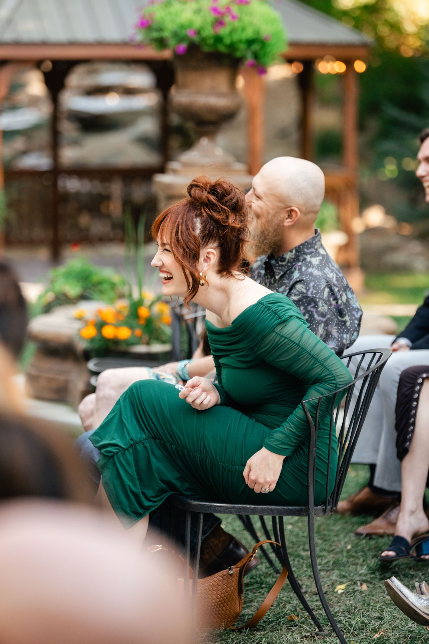 A couple shares a joyful moment together while sitting outdoors in a garden setting with greenery and flowers.