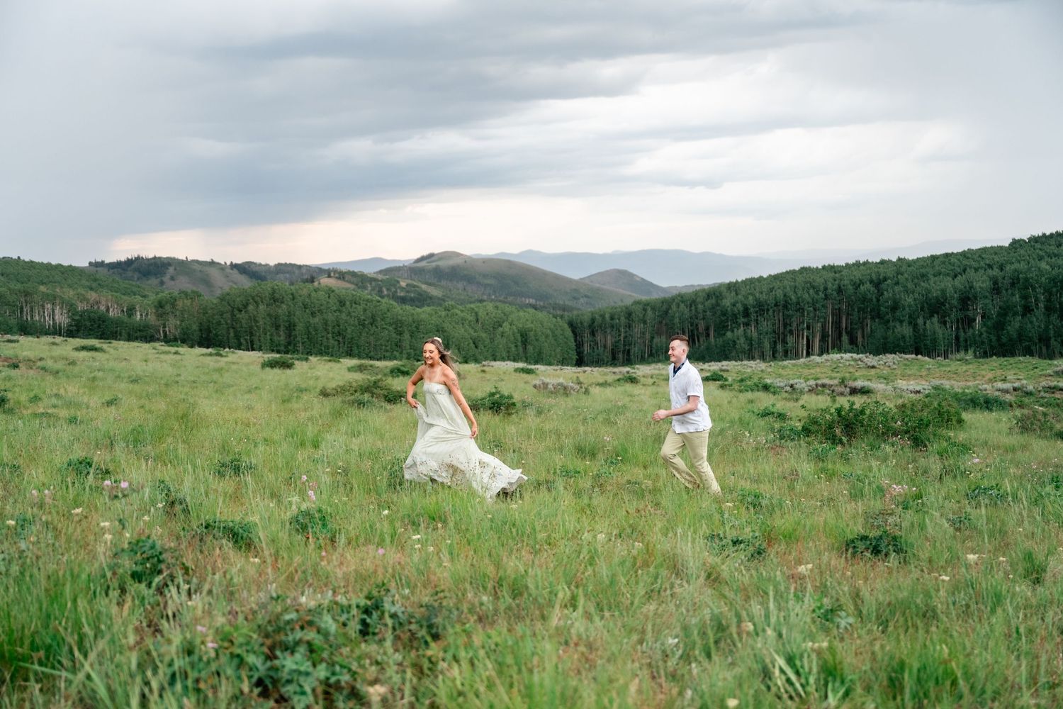 Two people in white attire run through a green meadow surrounded by mountains and forest on a cloudy day. Guardsman Pass engagement photos