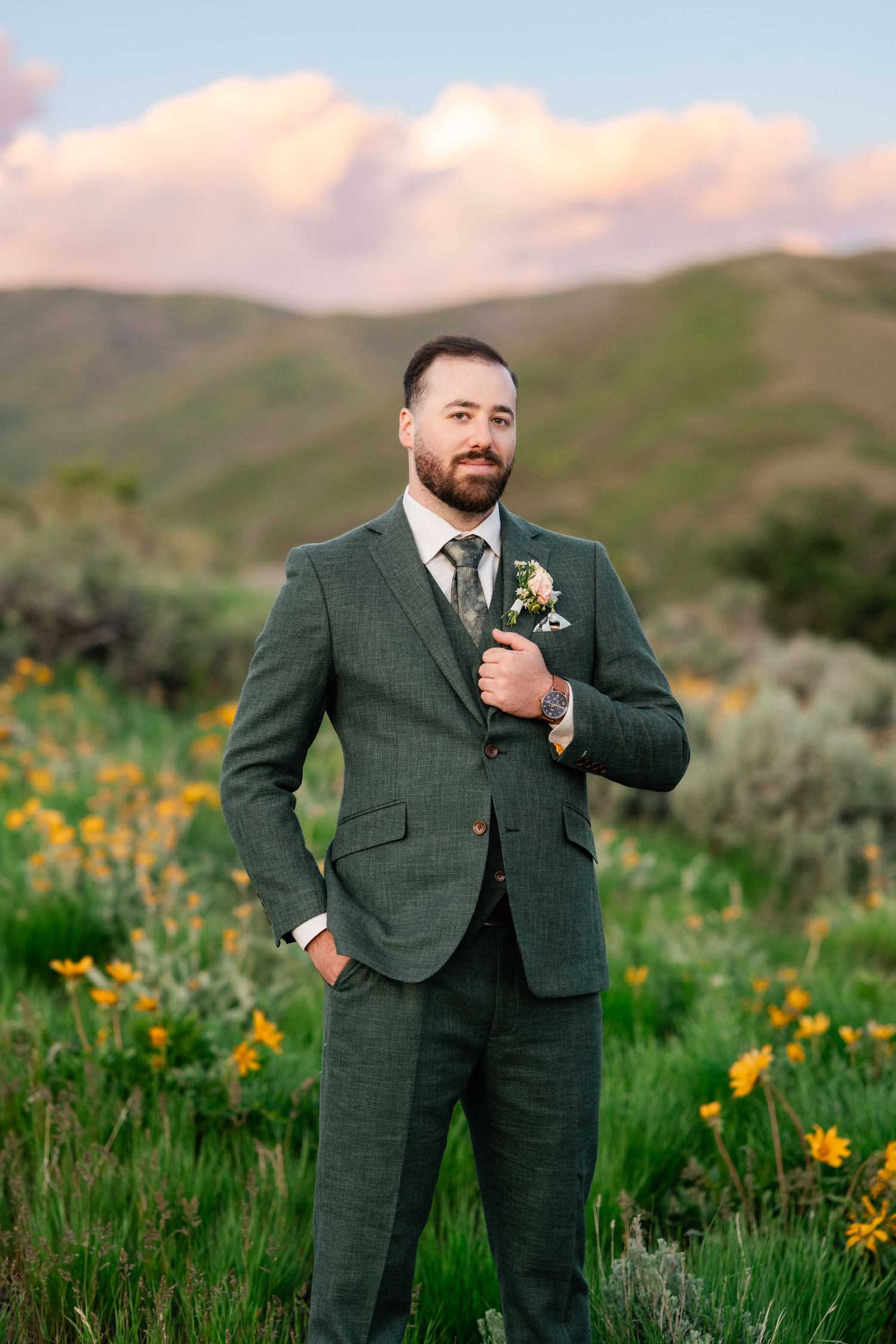 Groom in forest green suit stands among mountain wildflowers at sunset.