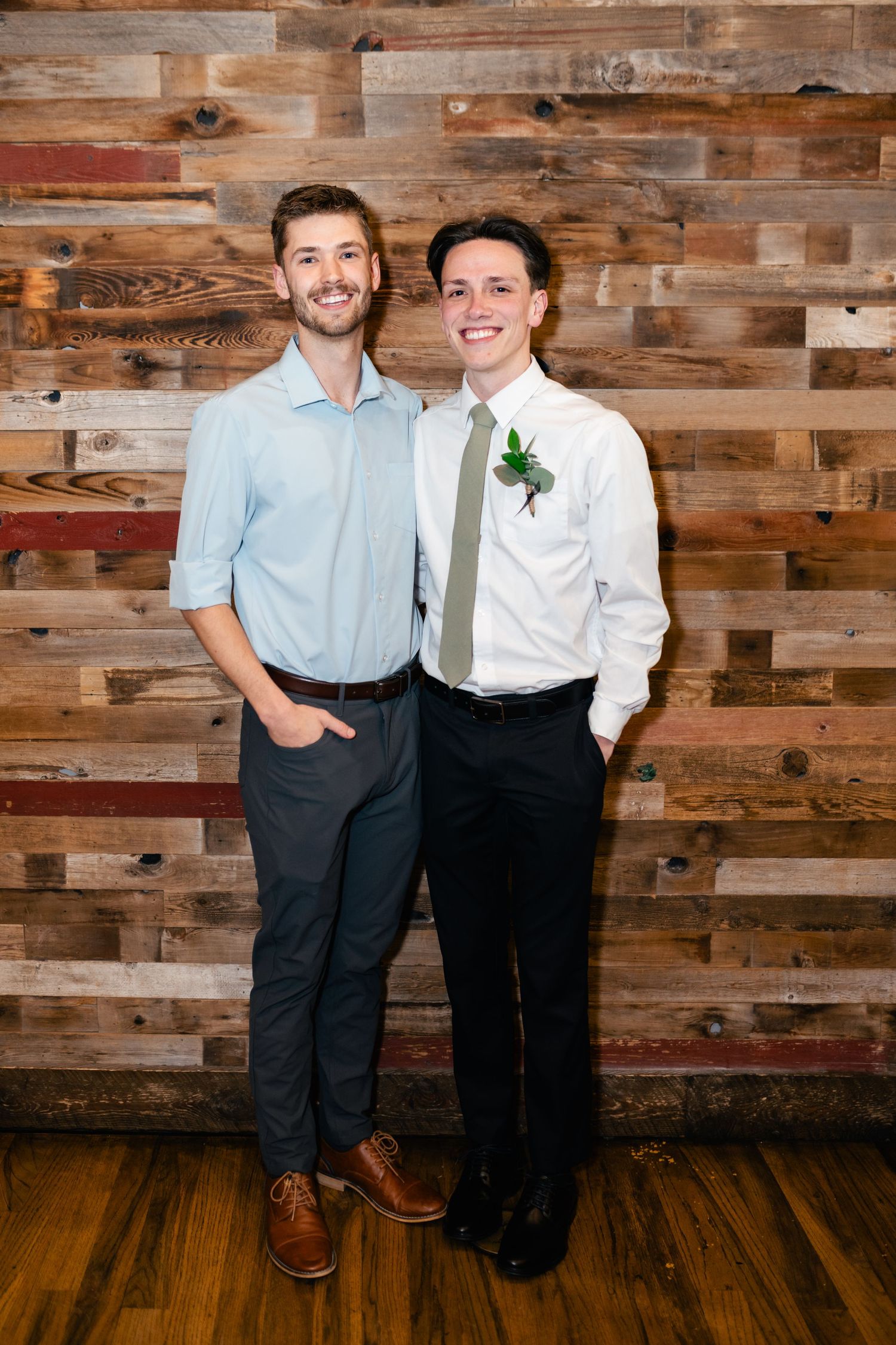 Two people in business casual attire stand together smiling in front of a rustic wooden plank wall.